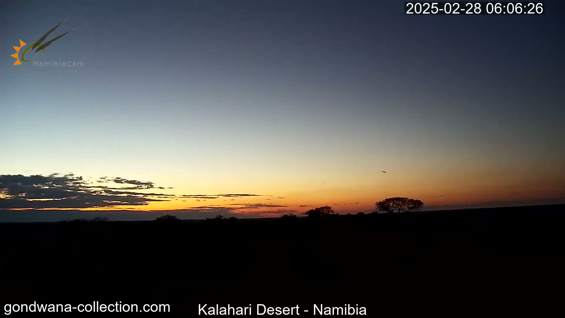 A sunset over a dark, flat landscape with a few silhouetted trees, under a mostly clear sky.