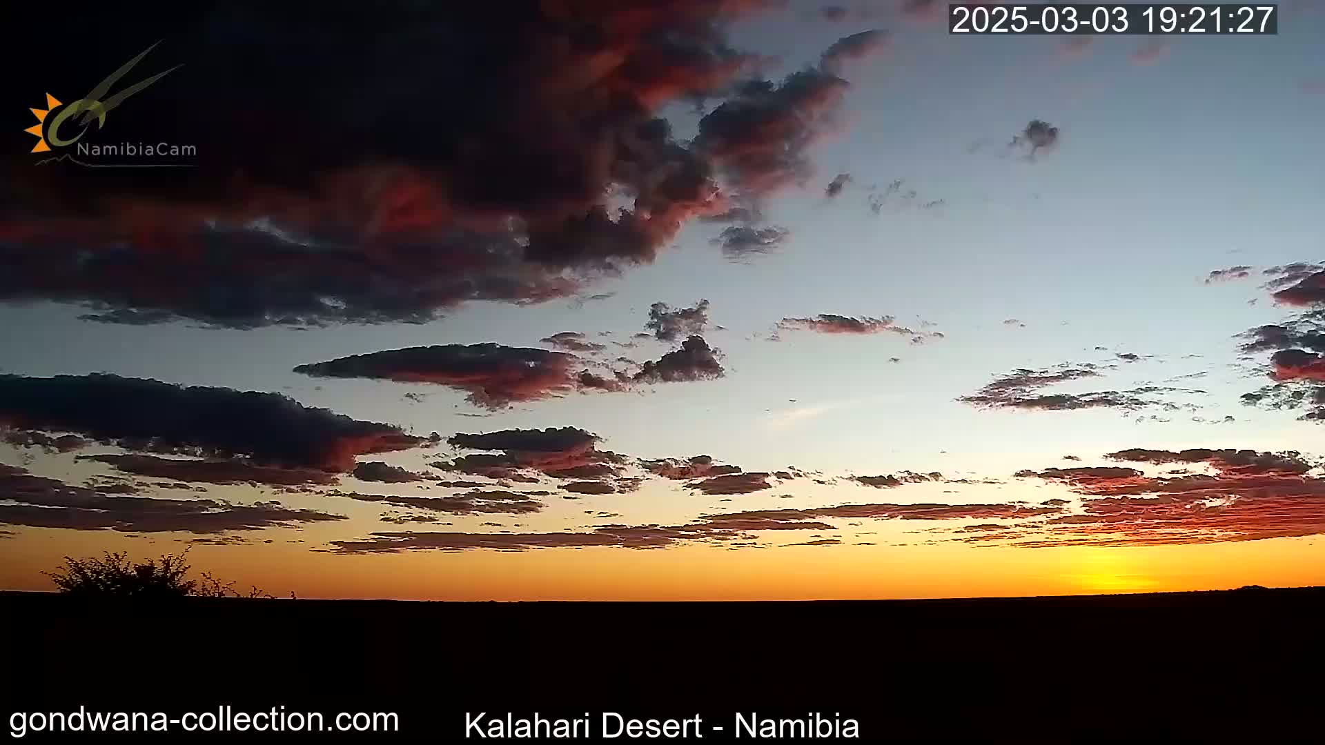 A fiery orange and red sunset fills the sky above a dark, flat landscape under a partly cloudy sky.