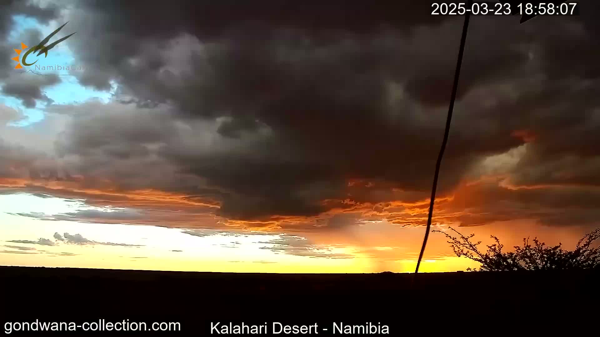 A dramatic sunset with dark, stormy clouds illuminates the Kalahari Desert sky in shades of orange and yellow.