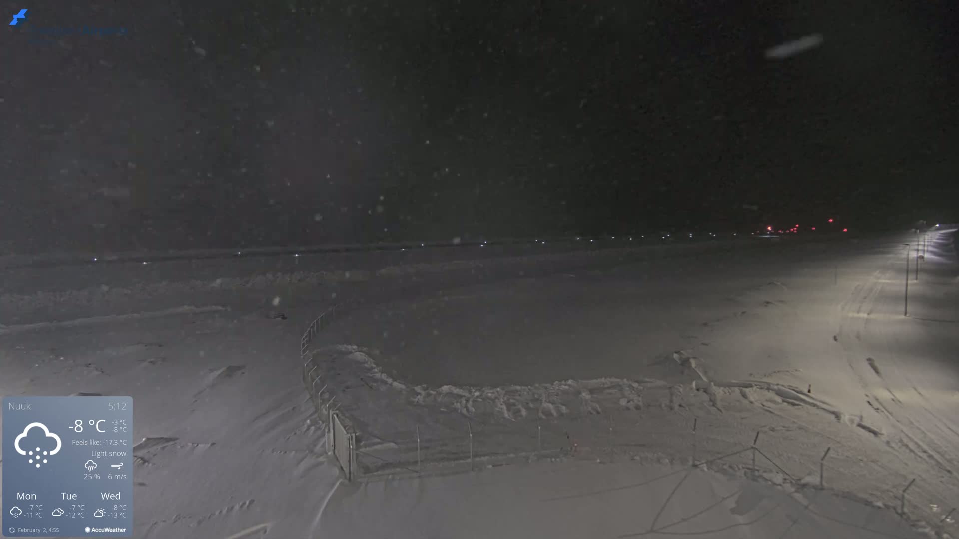 A snow-covered landscape at night, under light snowfall, is illuminated by streetlights along a road.