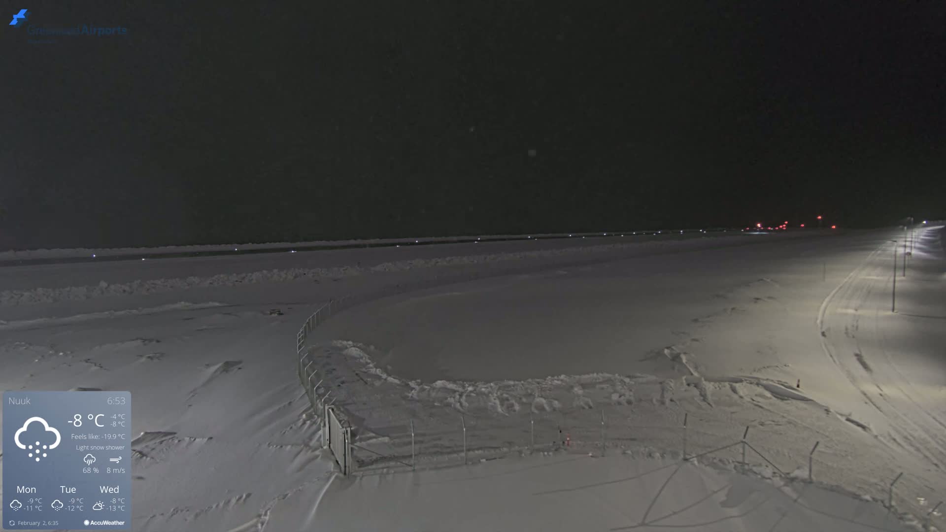 A snow-covered airfield is illuminated at night under a light snow shower.