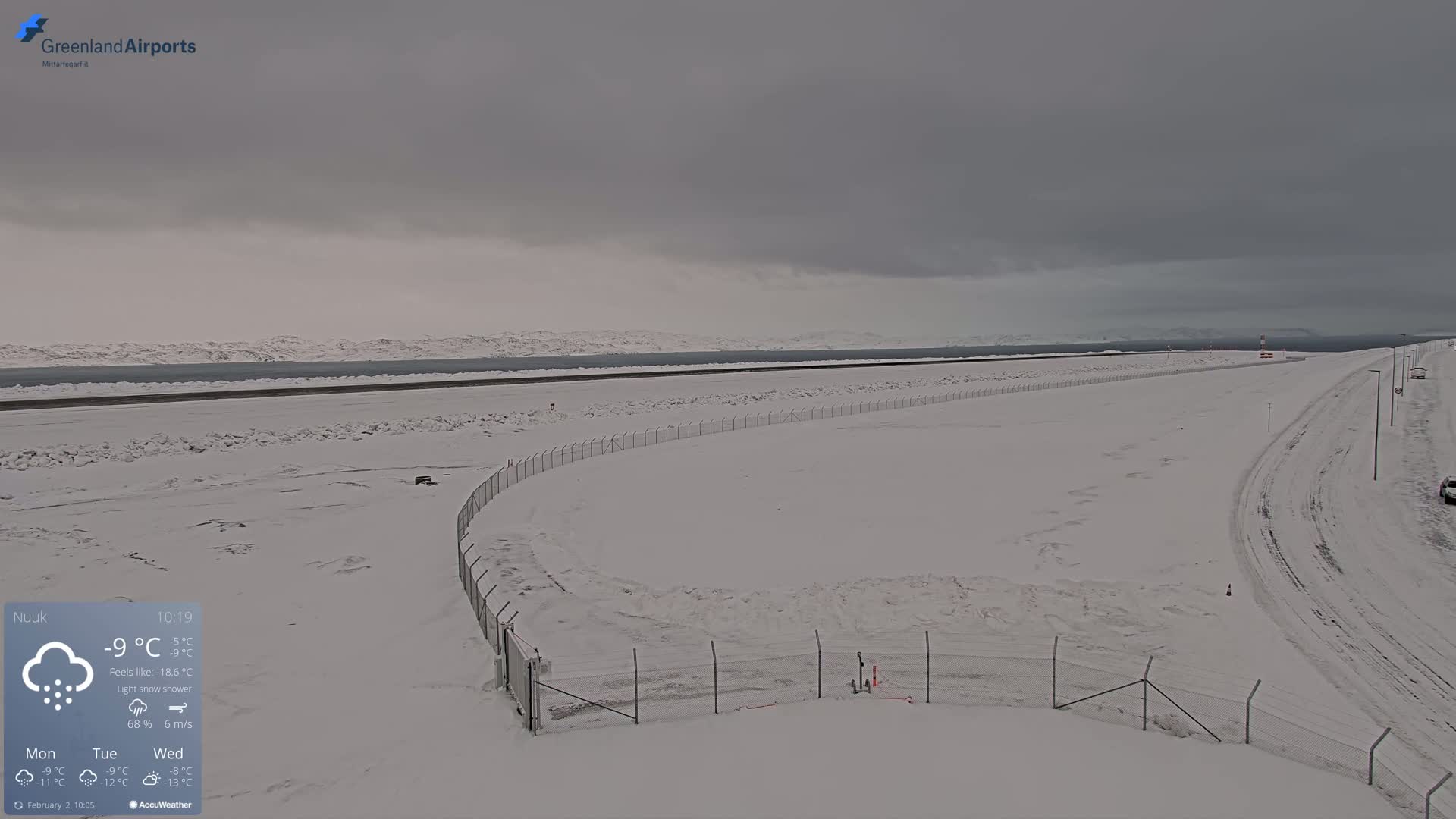 A snow-covered airport runway and surrounding area are visible under a cloudy, overcast sky with a light snow shower.