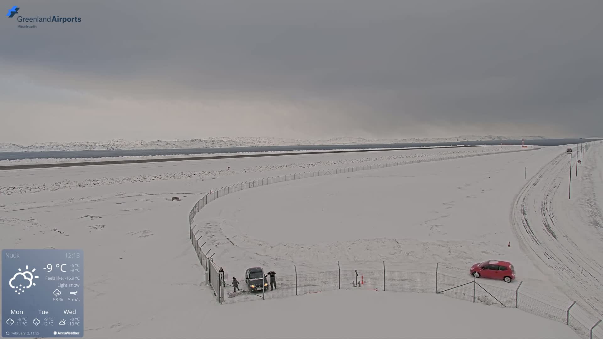 A snow-covered airport runway and surrounding area with a few vehicles, under a light snow sky.