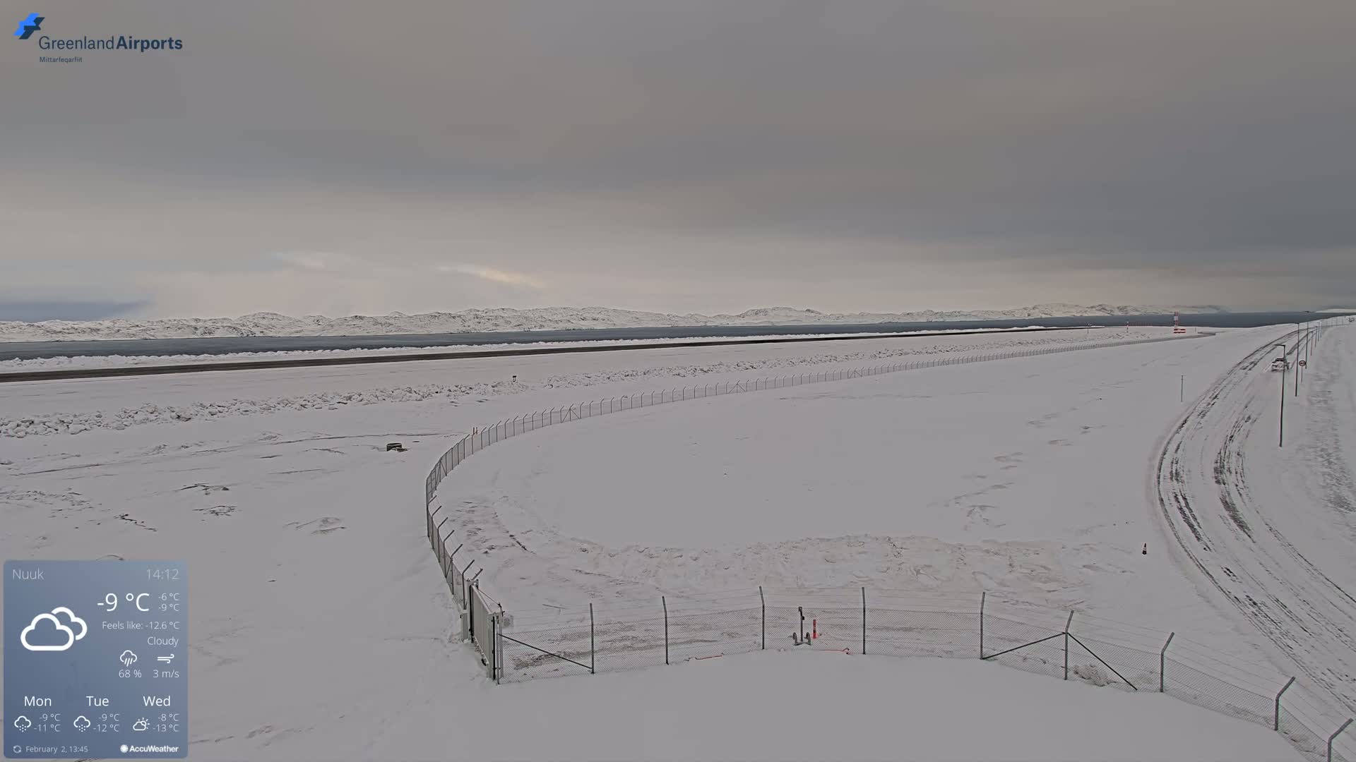 A snow-covered airport runway and surrounding area are visible under a cloudy sky.