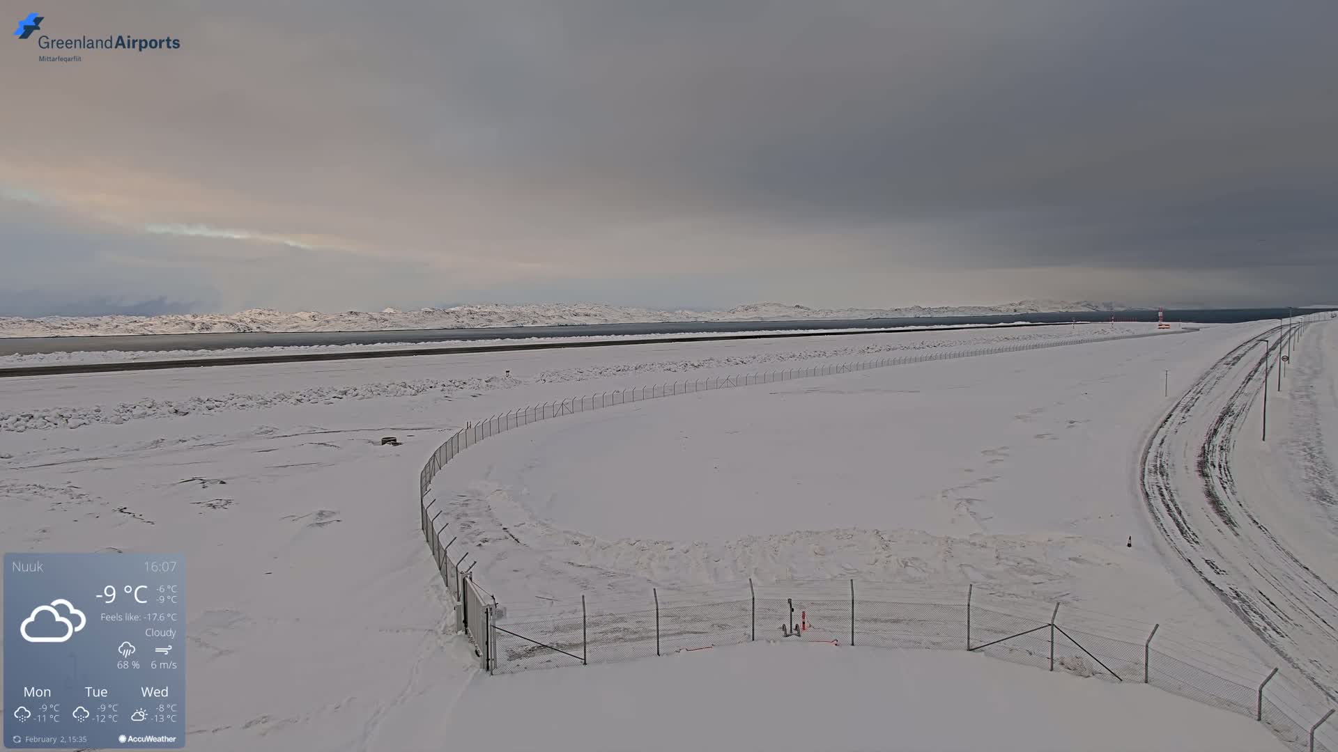 A snow-covered airport tarmac and surrounding area under a cloudy sky, with a body of water and distant mountains visible in the background.