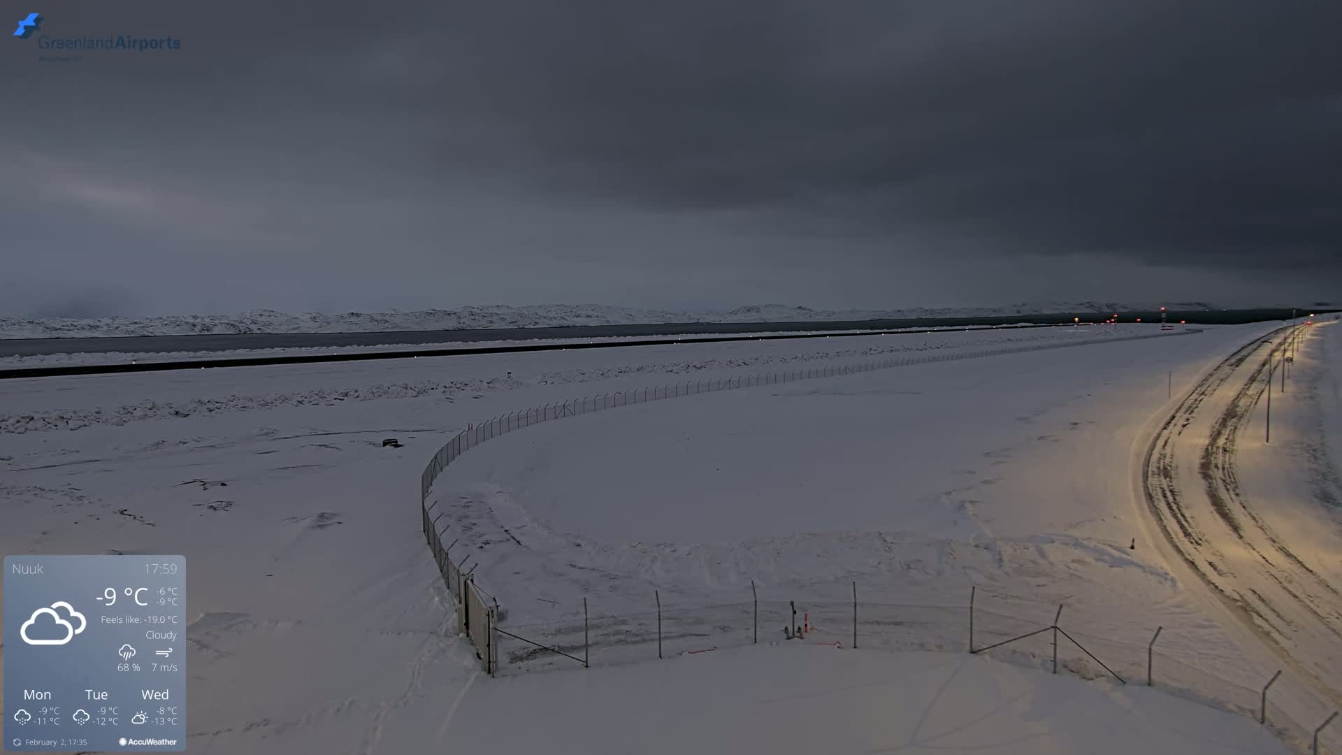 A snow-covered landscape with a road and fence under a cloudy, dark sky.