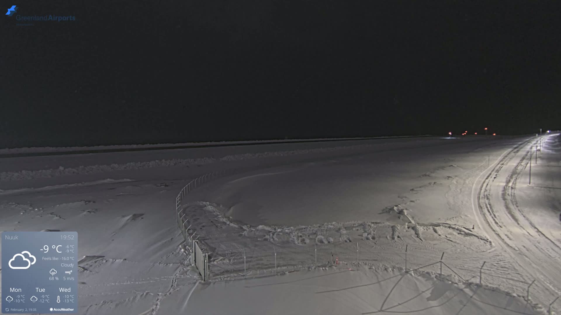 A snow-covered, dark landscape at night, illuminated by streetlights, shows a road and fence under cloudy skies.