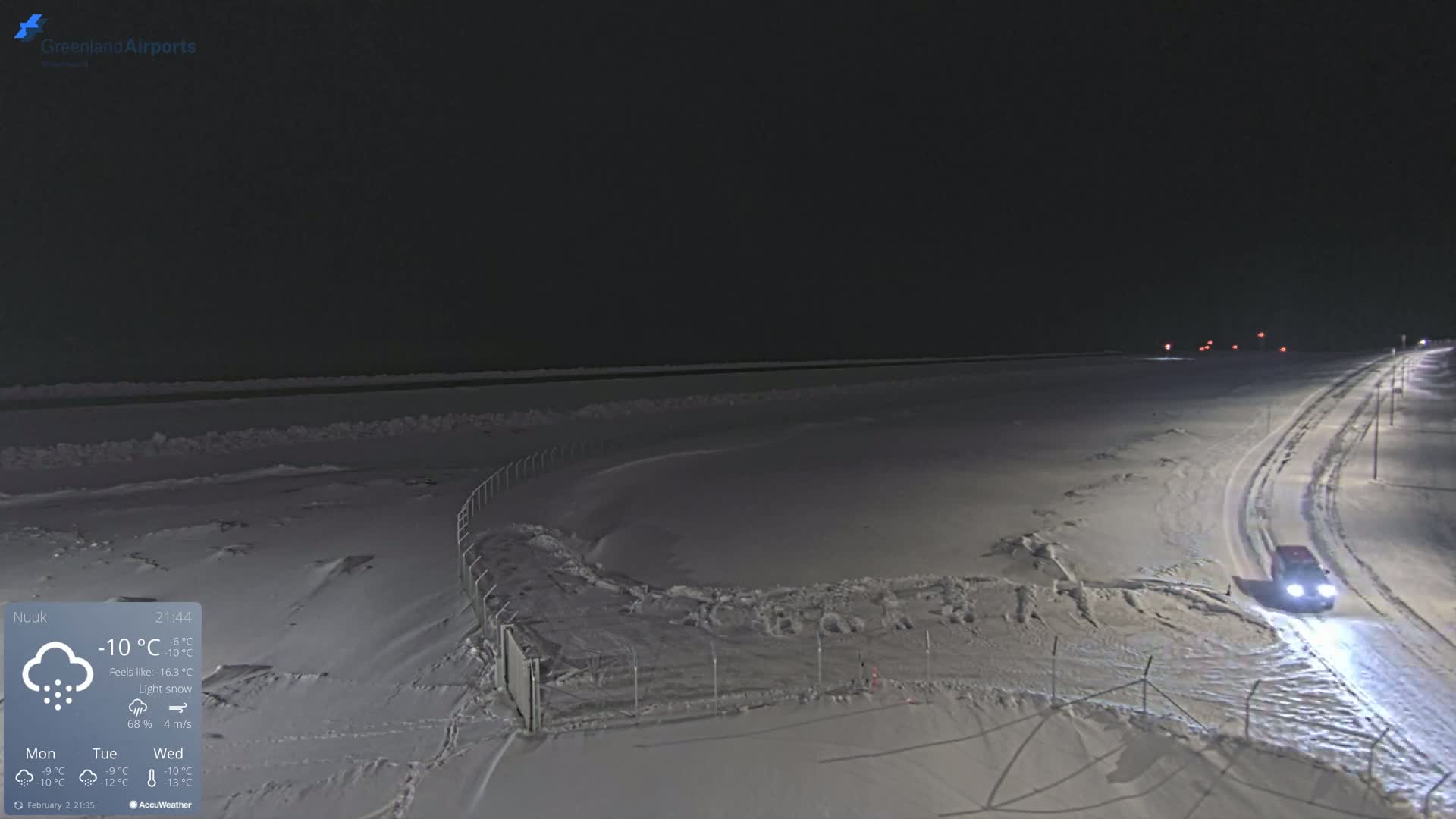 A snow-covered landscape at night under light snowfall, with a vehicle driving on a road and a fenced area in the foreground.