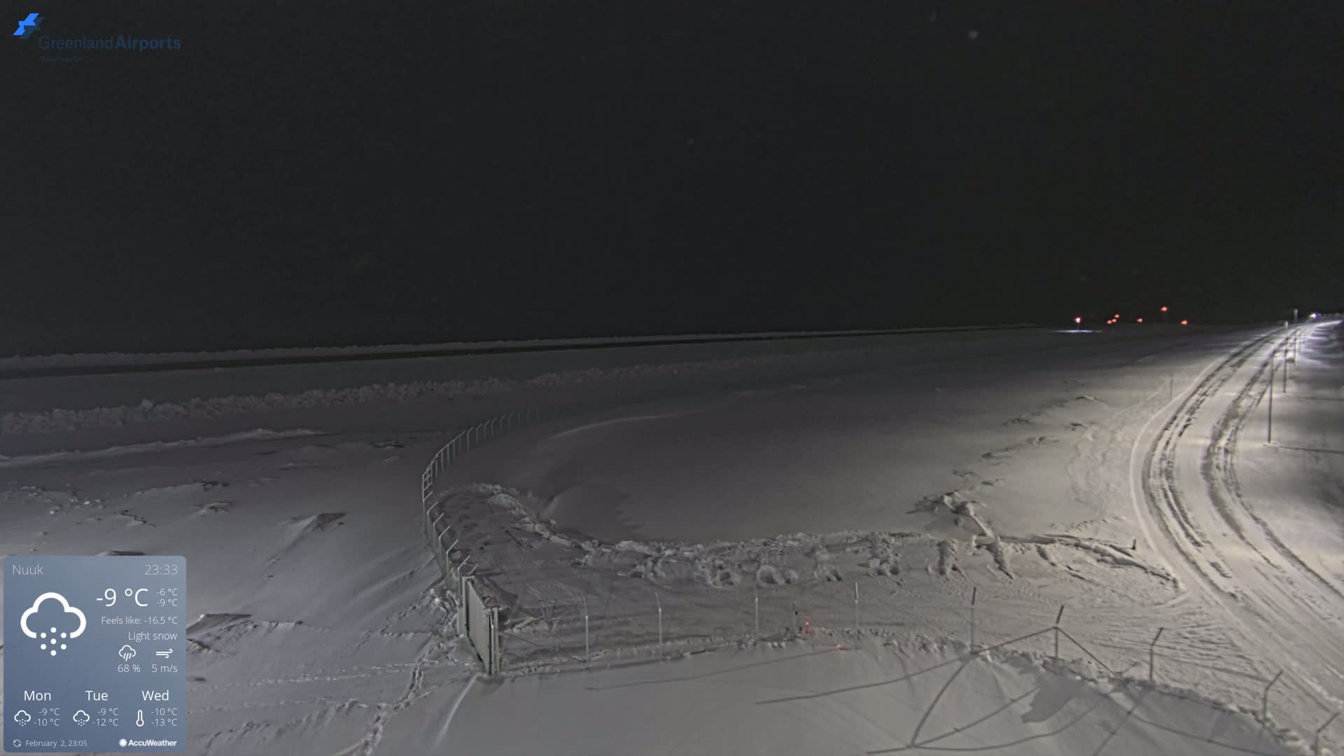 A snow-covered, dark landscape at night with a road and fence visible under light snow.