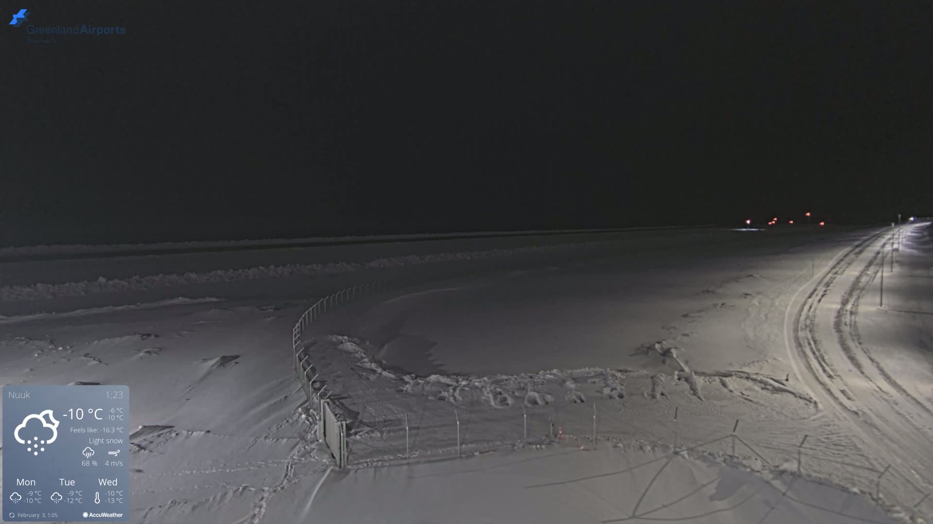 A snow-covered landscape at night with a road and fence visible under light snow.