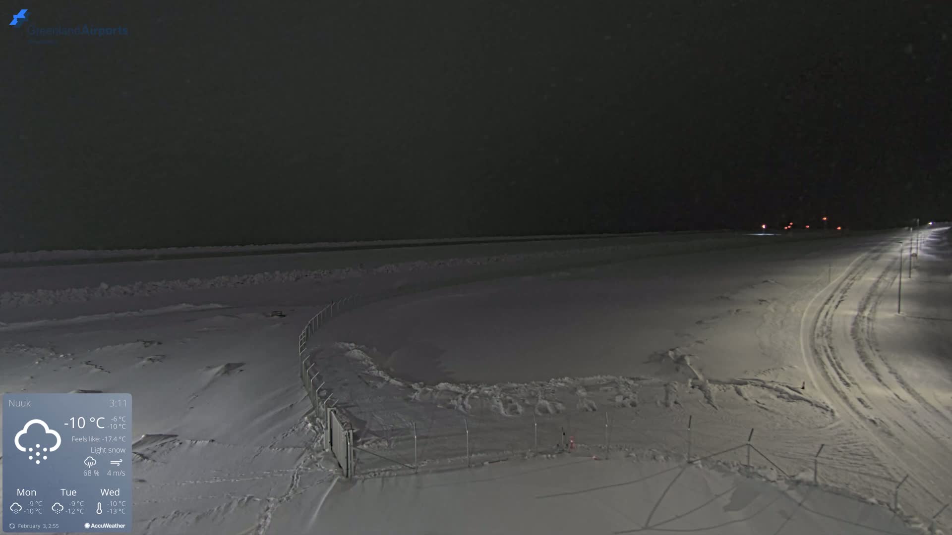 A snow-covered landscape at night, under light snow with a road curving into the distance, lit by streetlights.