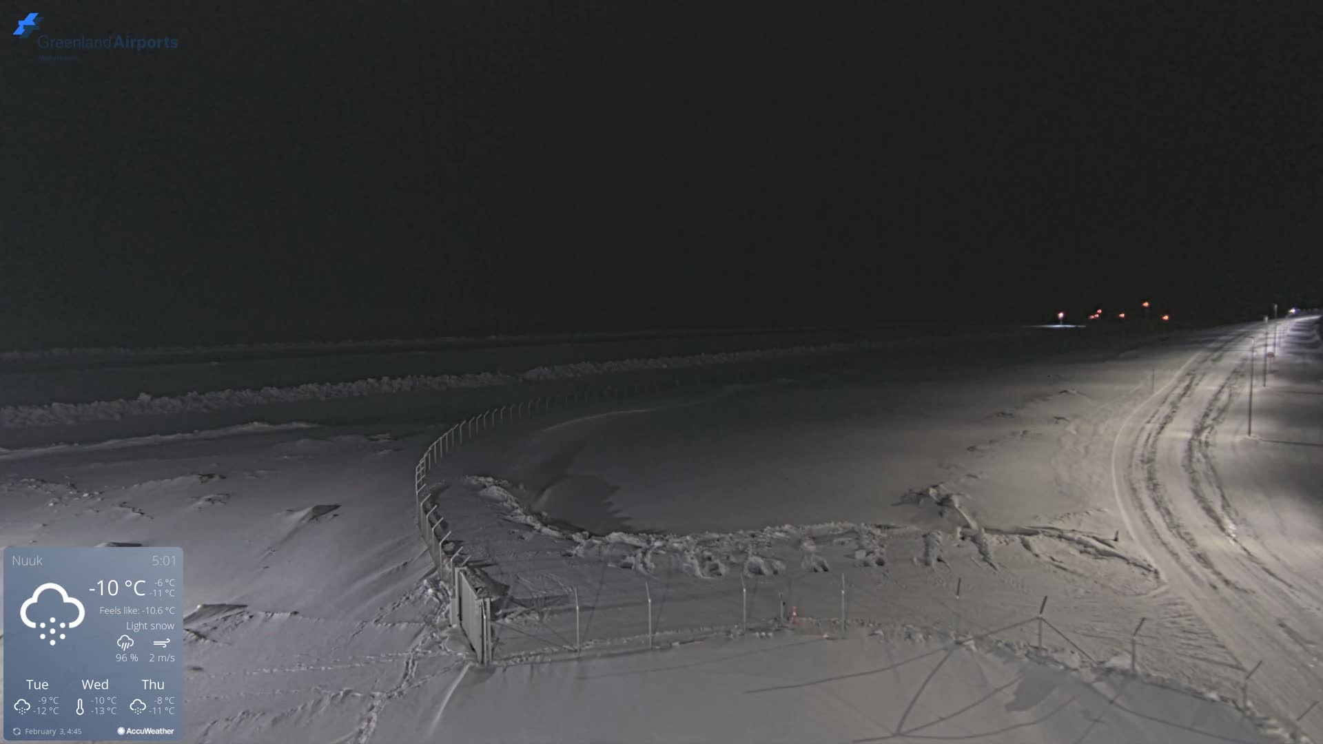 A snow-covered, nighttime landscape features a road with tire tracks, a fence surrounding a snow-filled area, and a few distant lights under light snow conditions.