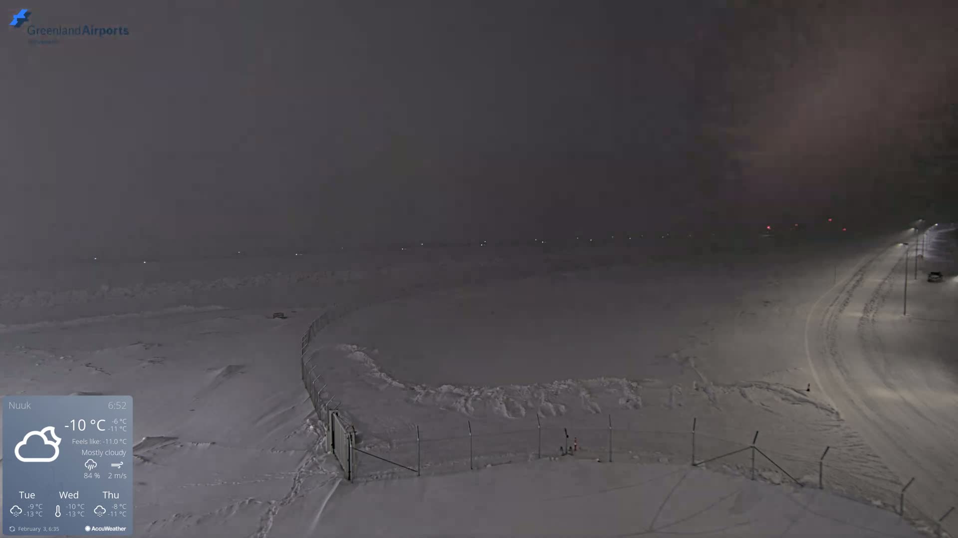 A snow-covered landscape at night under mostly cloudy skies, with a fence and a road visible.