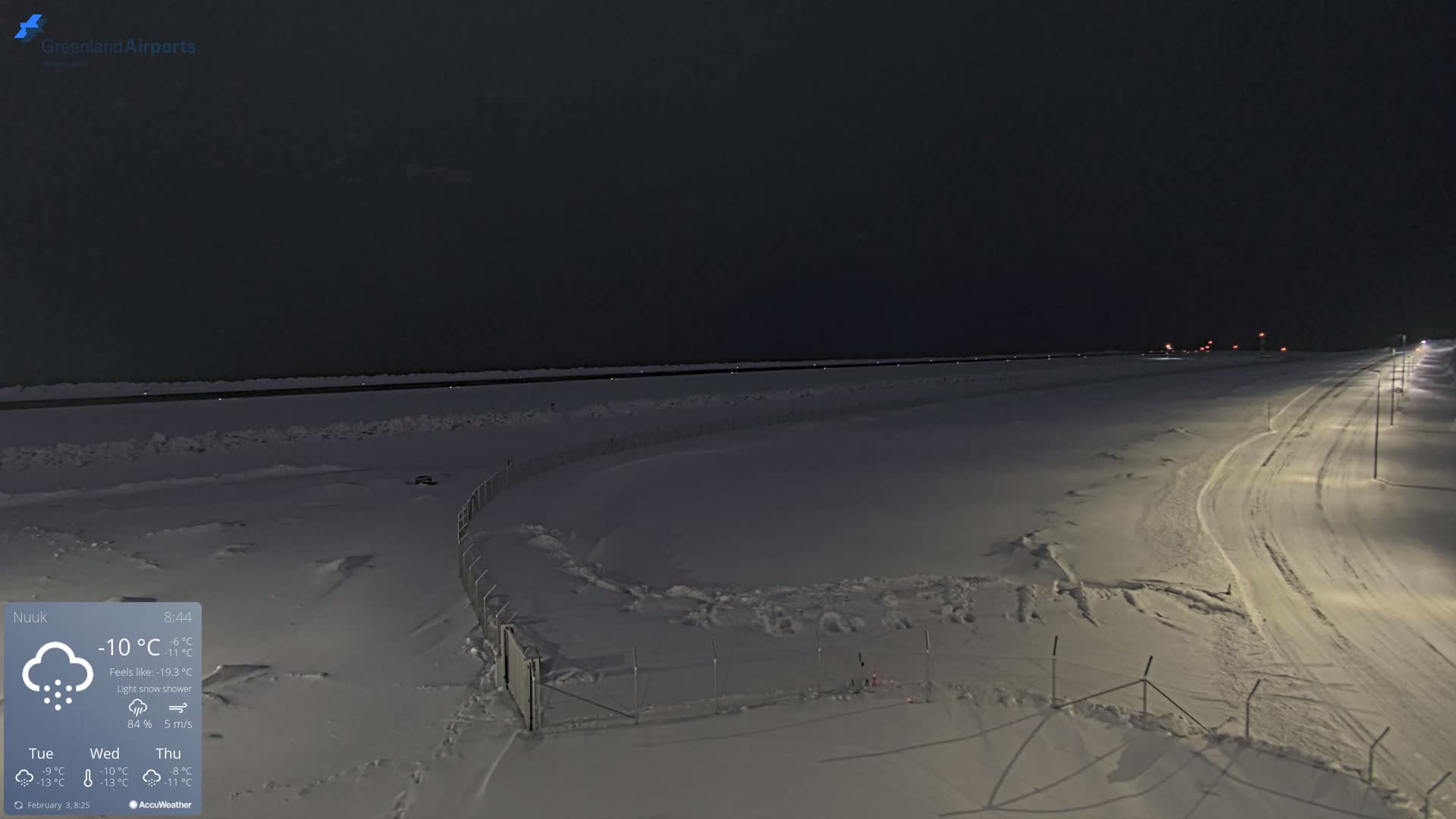 A snow-covered, dimly lit airport runway and adjacent road are visible under a dark, night sky with a light snow shower.