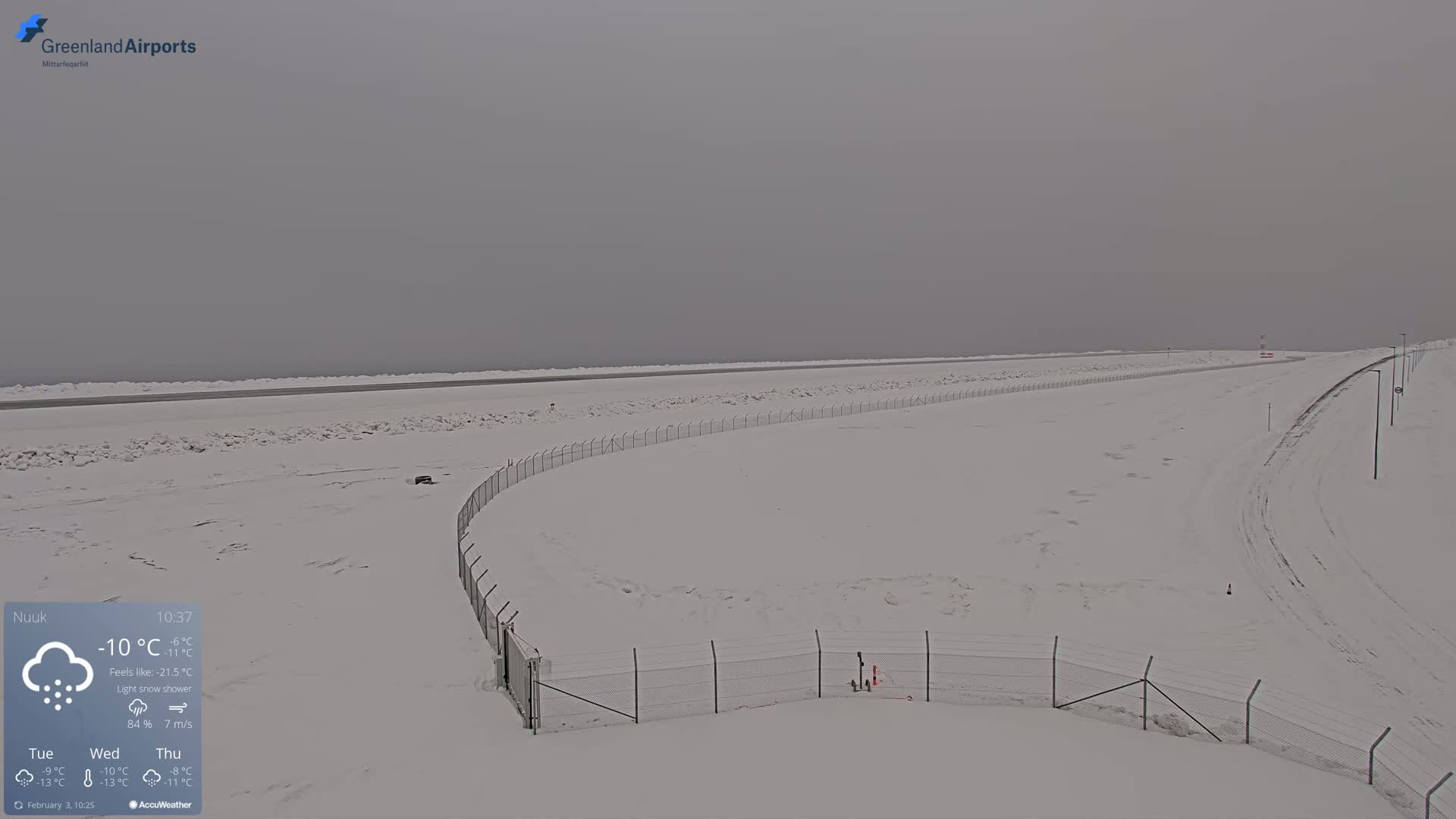 A snow-covered airport runway and surrounding area are visible under a light snow shower in overcast conditions.