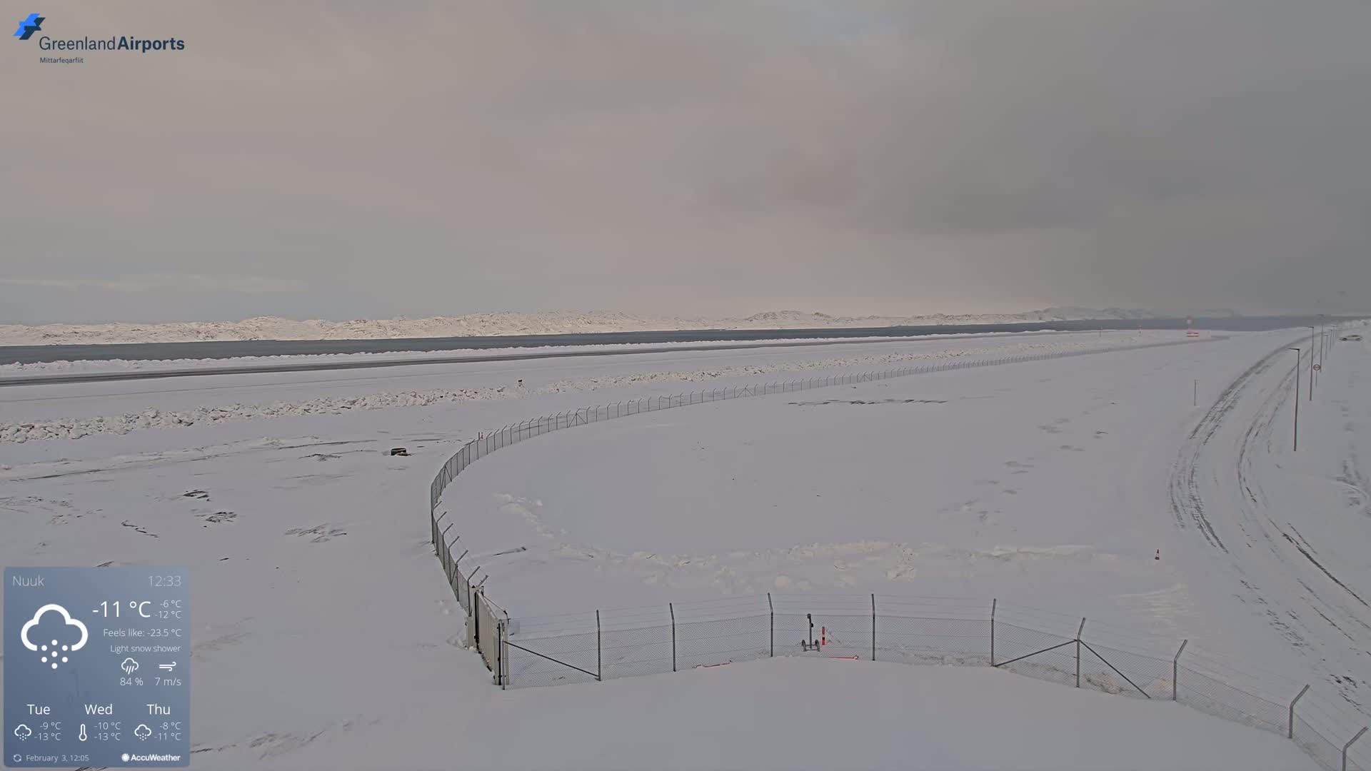 A snow-covered airport runway stretches toward a distant, snow-dusted mountain range under a light snow shower.