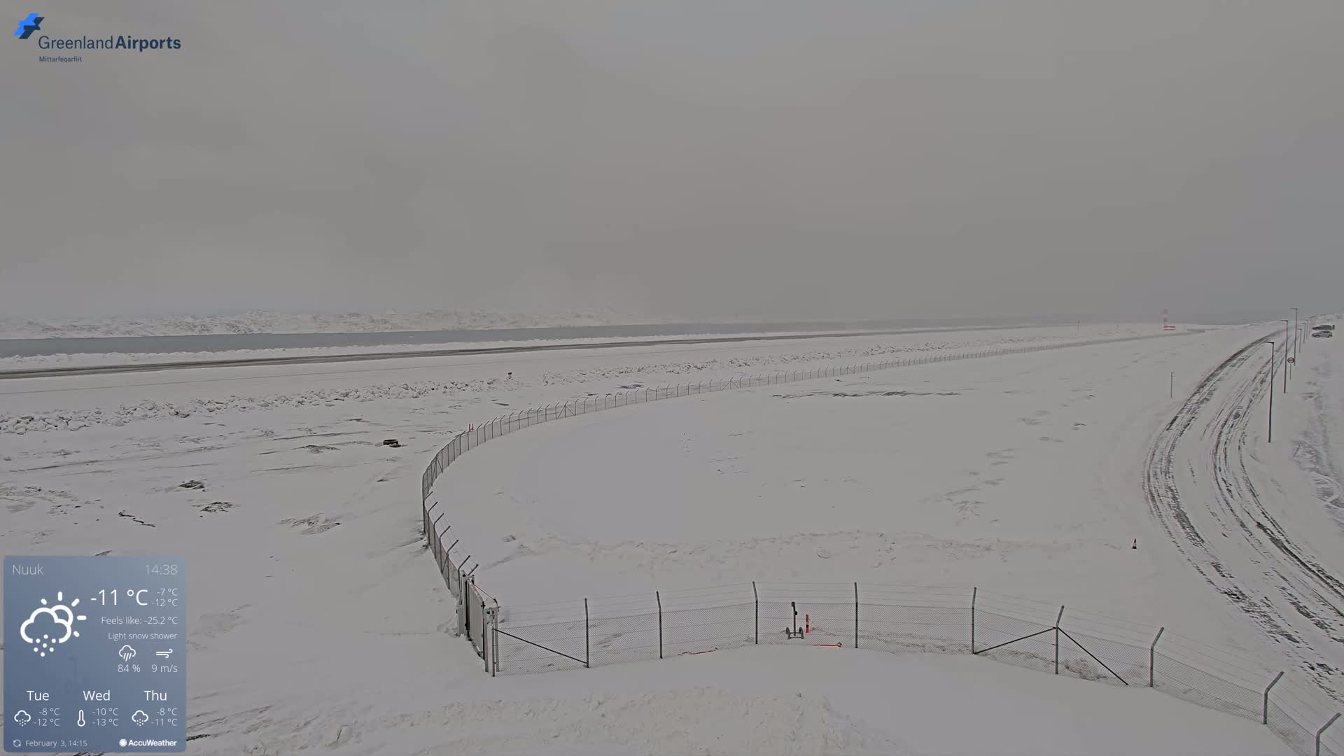 A snow-covered airfield with a chain-link fence and a road, under a light snow shower, overlooks a frozen body of water.
