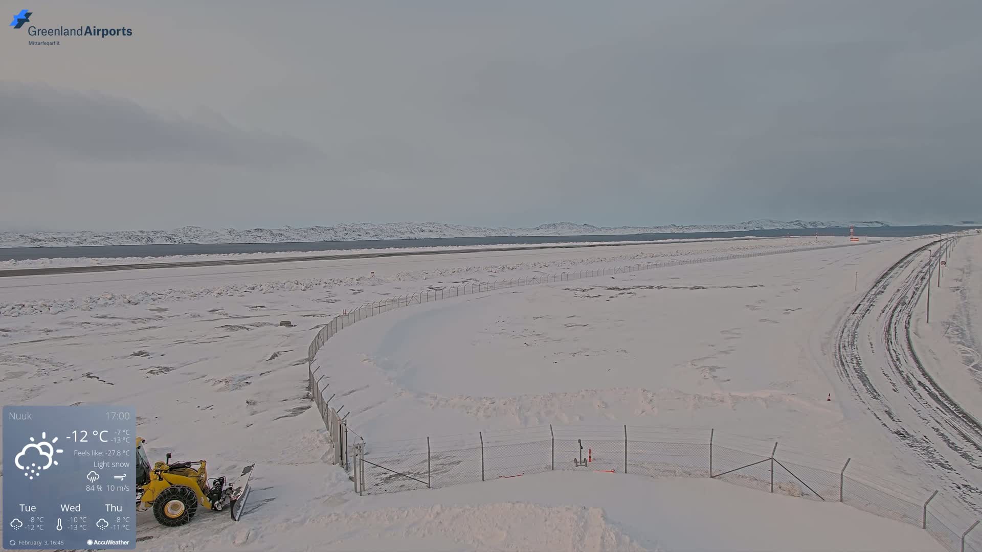 A snow-covered airport with a small yellow snowplow is seen under a light snow with overcast skies and a frozen body of water and distant mountains in the background.