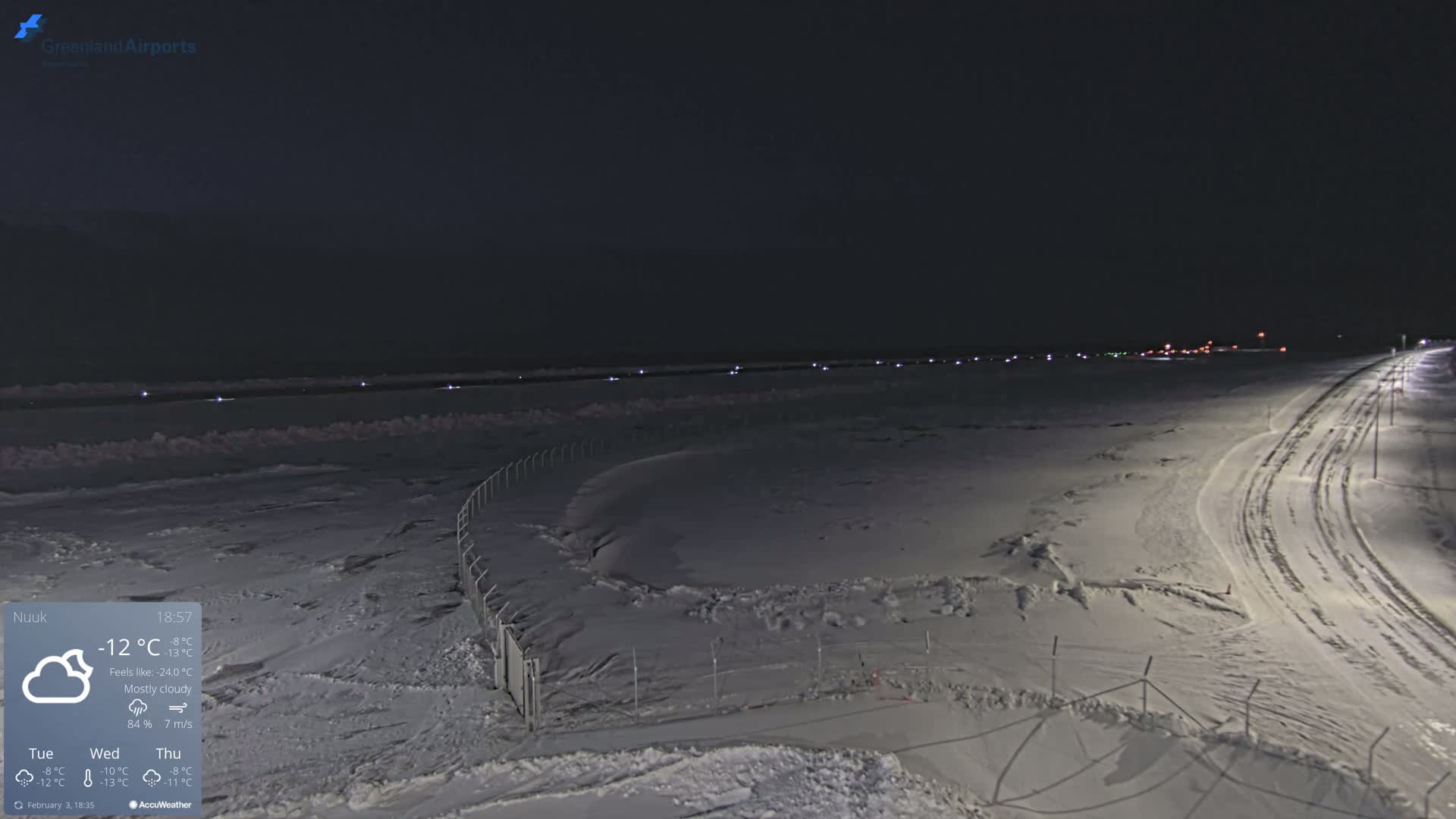 A snow-covered airport runway at night under mostly cloudy skies, with lights illuminating the runway and surrounding area.