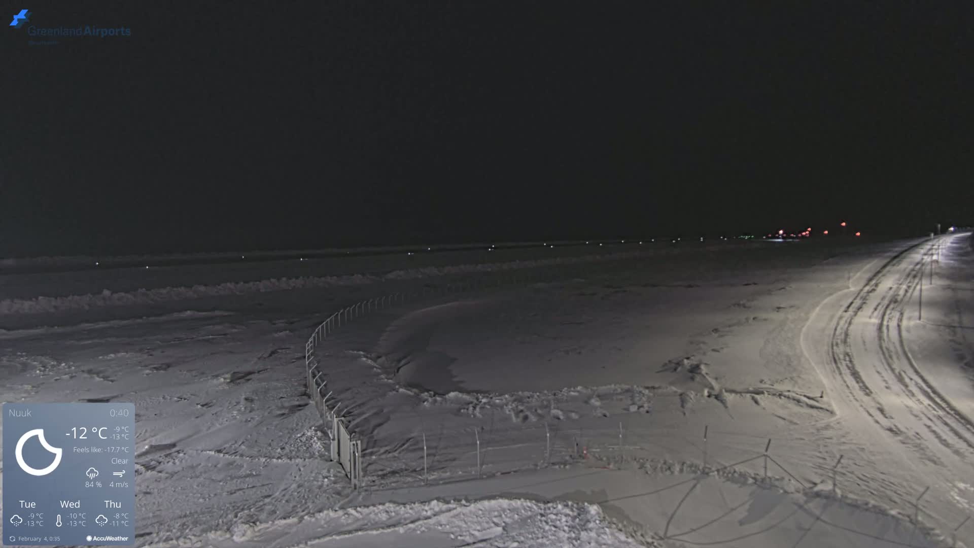 A snow-covered landscape at night, illuminated by distant lights and featuring a fence and a road with tire tracks.