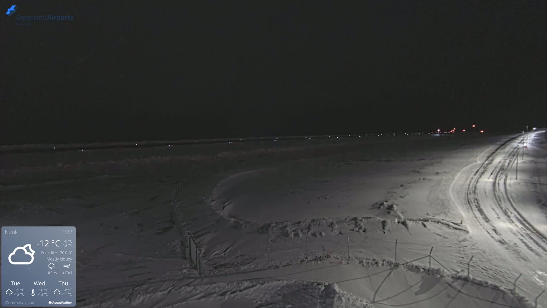 A snow-covered landscape at night under mostly cloudy skies, with a road curving into the distance, illuminated by lights.
