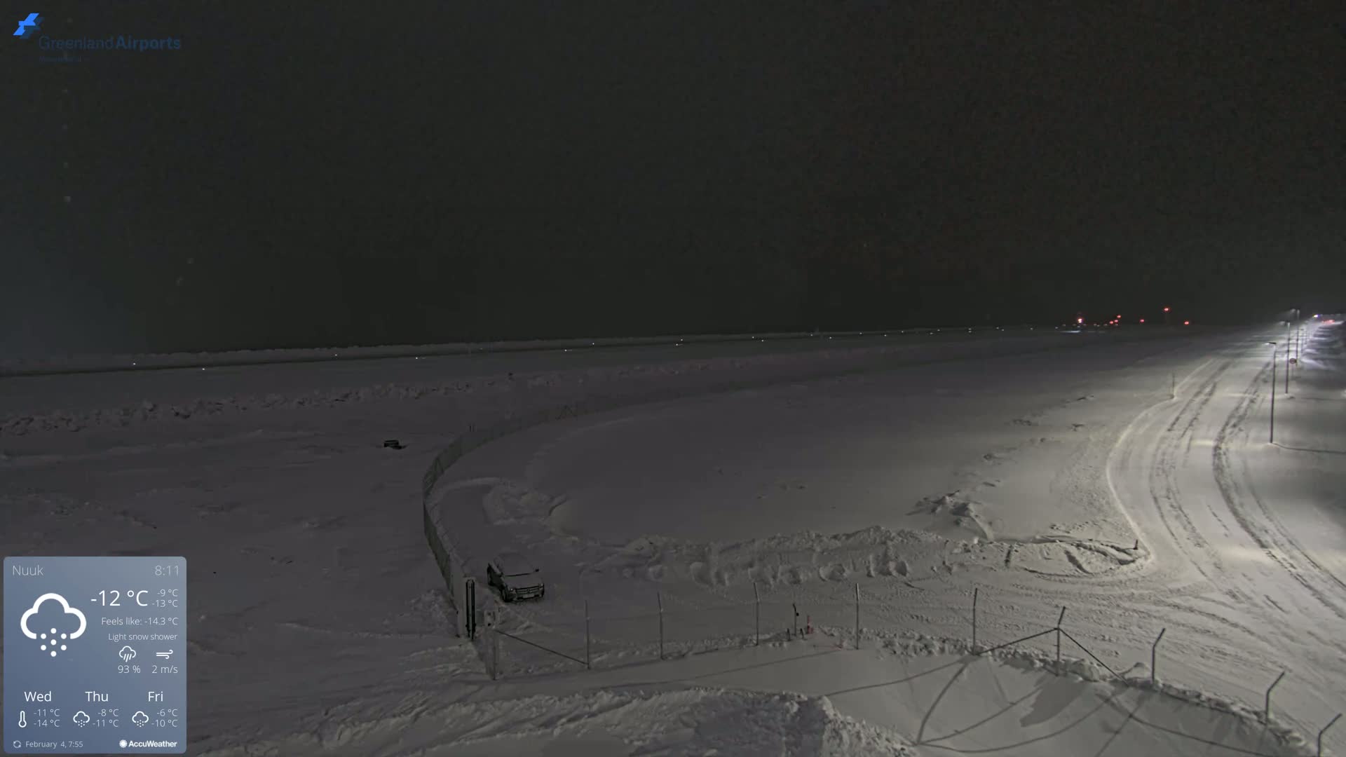 A snow-covered airport runway and surrounding area at night during a light snow shower.