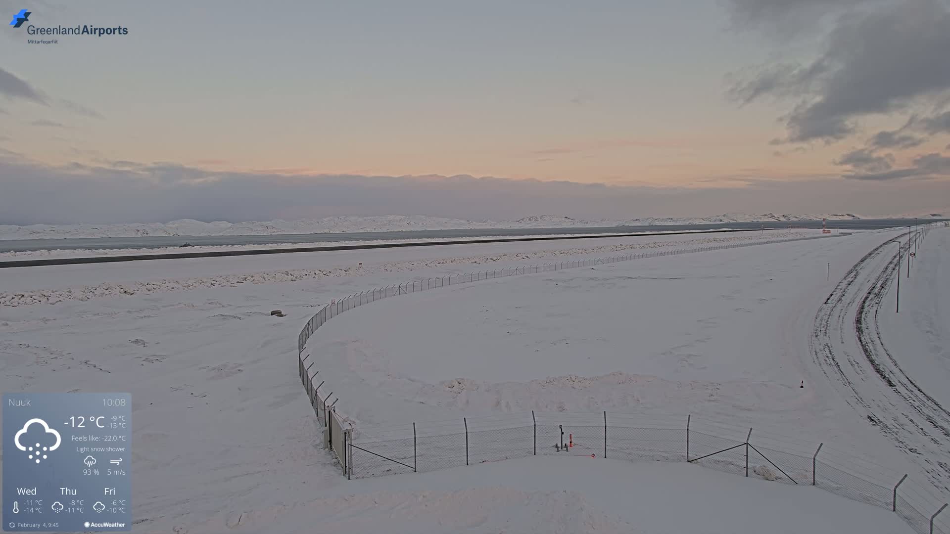 A snow-covered airport runway extends to a body of water under a light snow shower and pale pink sky.