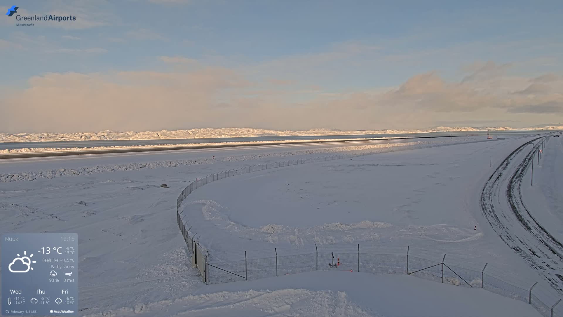 A snow-covered airport runway and surrounding area are visible under a partly sunny sky, with mountains in the background.