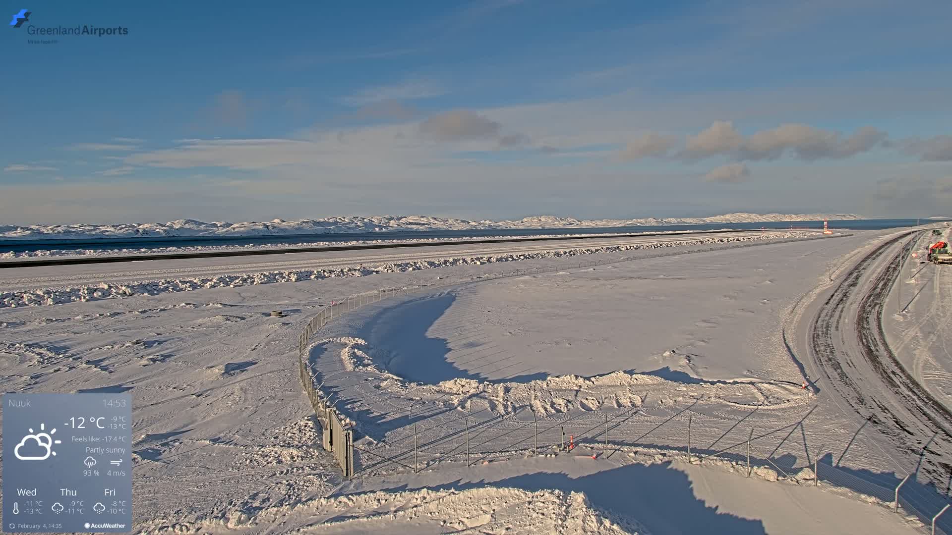 A snow-covered airport runway and surrounding area are visible, with partly sunny skies and mountains in the distance.