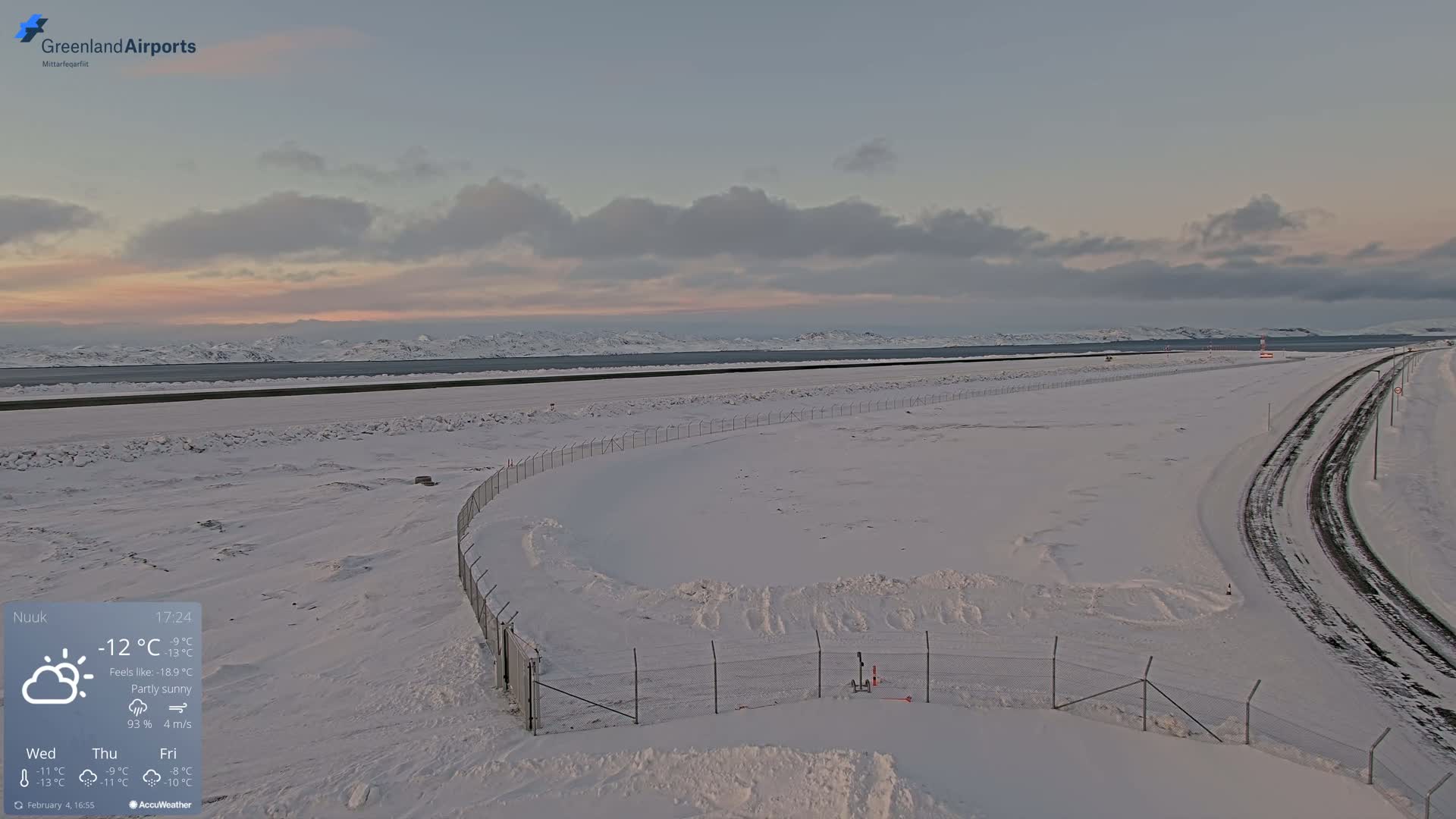 A snow-covered airport runway and surrounding area are visible under a partly sunny sky.