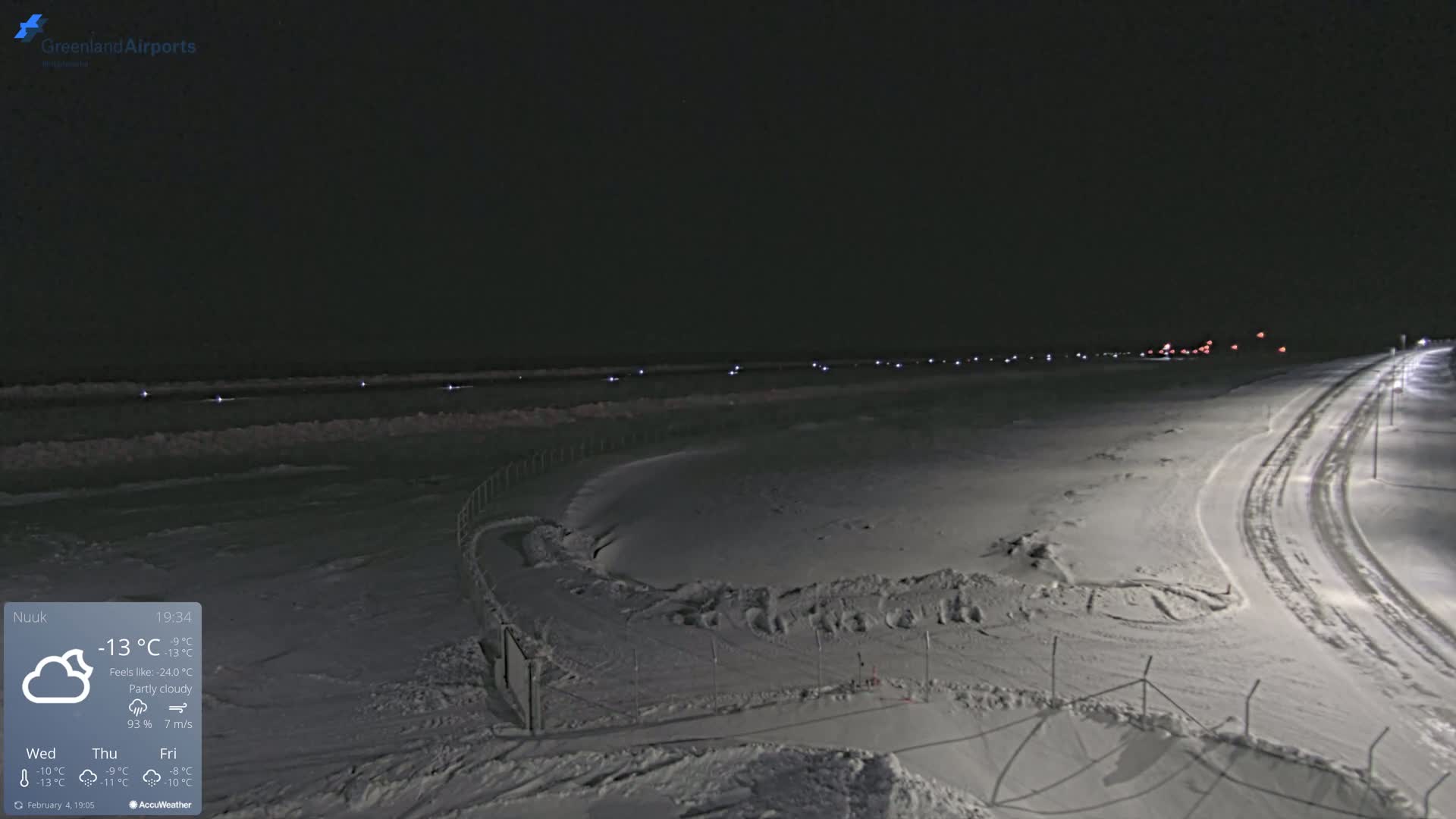 A snow-covered landscape at night under partly cloudy skies, showing a road curving toward distant lights, with a fence and faint lights visible along a body of water in the background.