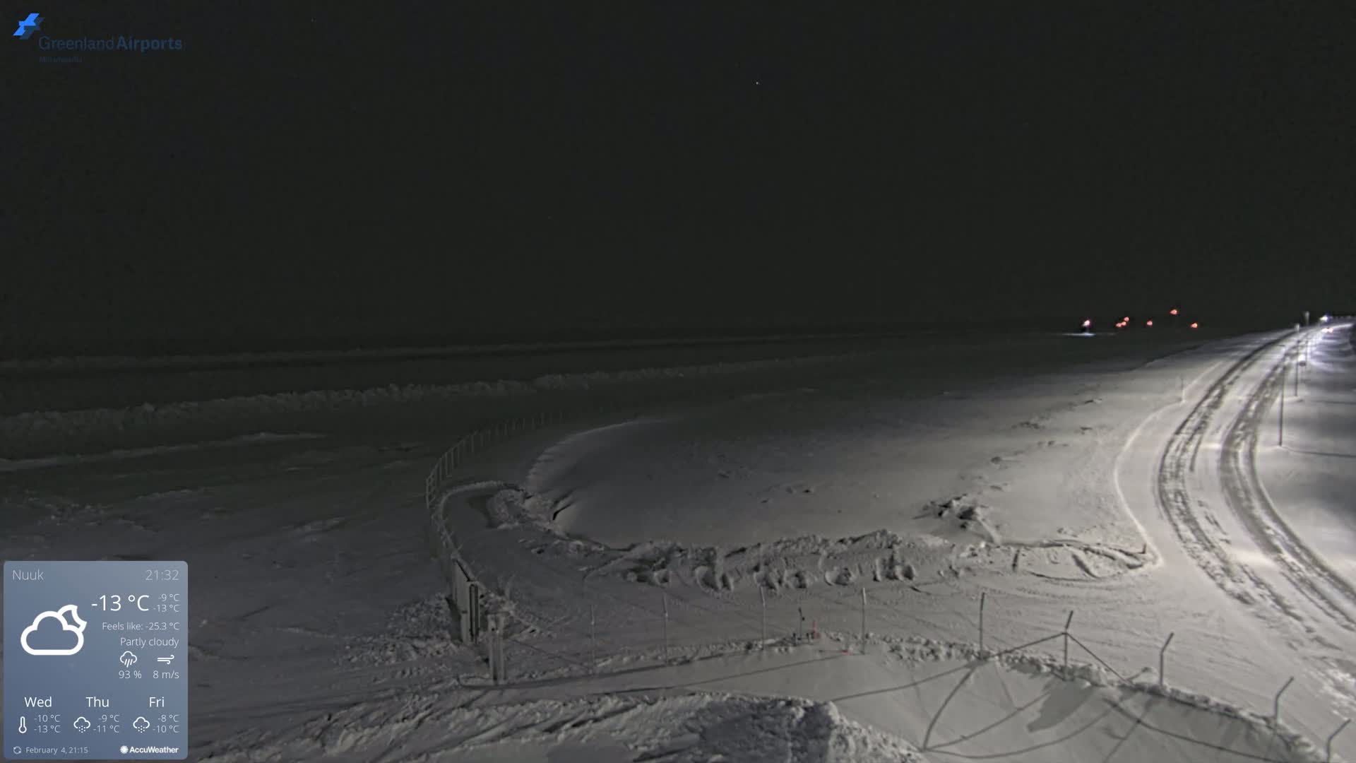 A snow-covered landscape at night, under partly cloudy skies, shows a road with tire tracks and a fenced-in area.