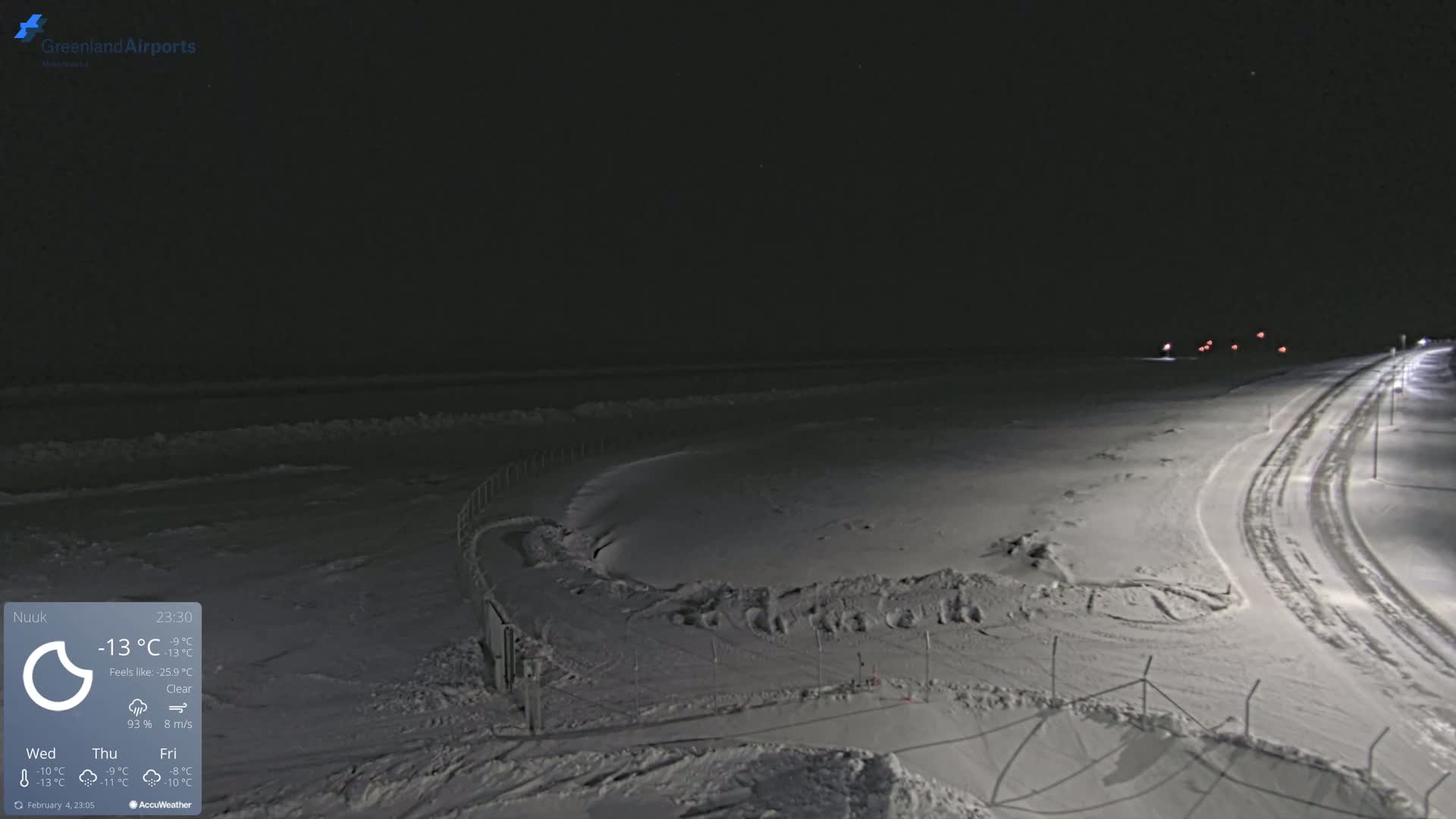A snow-covered, dark nighttime scene shows a road curving toward a body of water partially covered in ice under a clear sky.