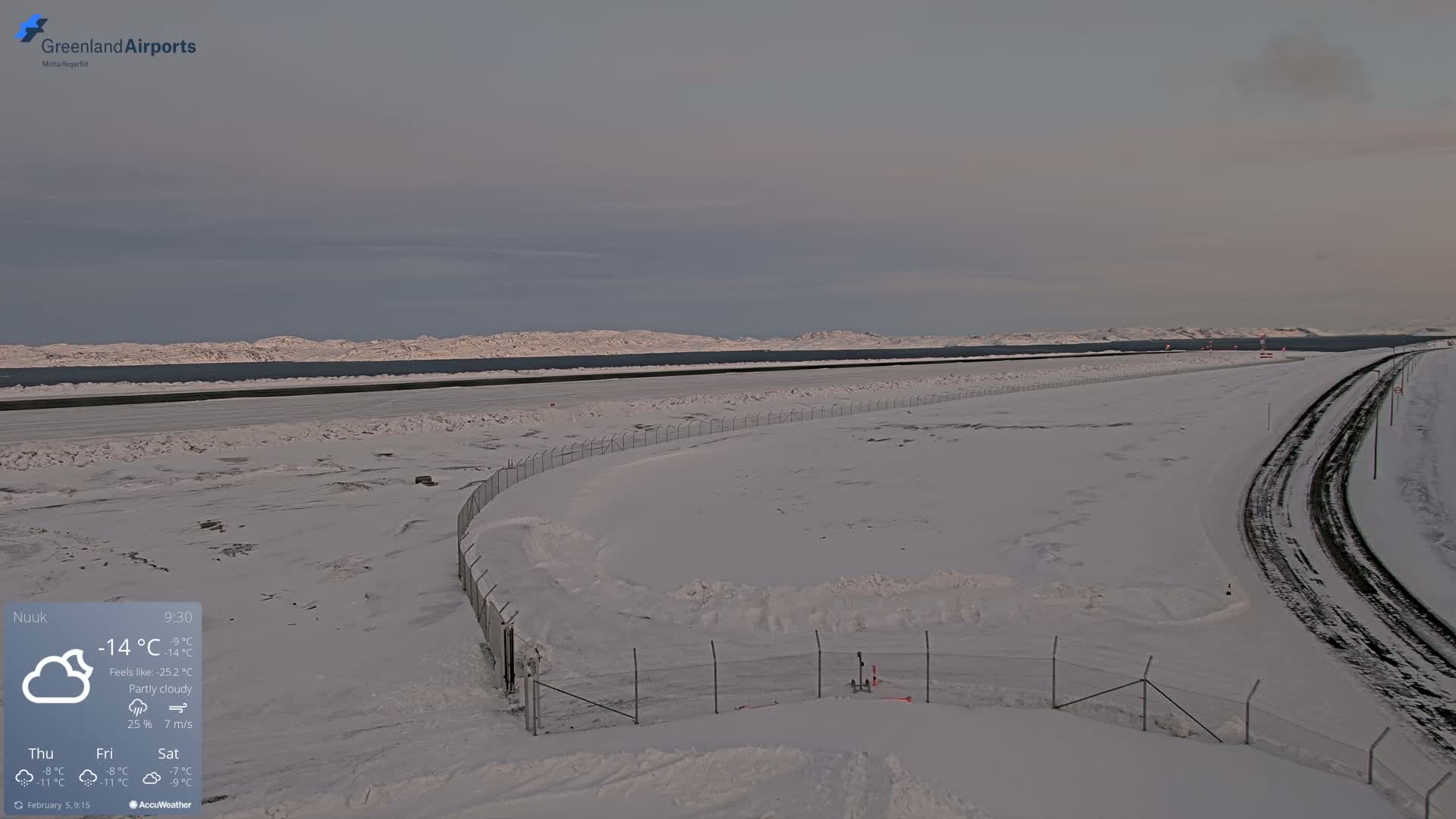 A snow-covered airport runway and road extend toward a frozen body of water and distant, snow-capped mountains under a partly cloudy sky.