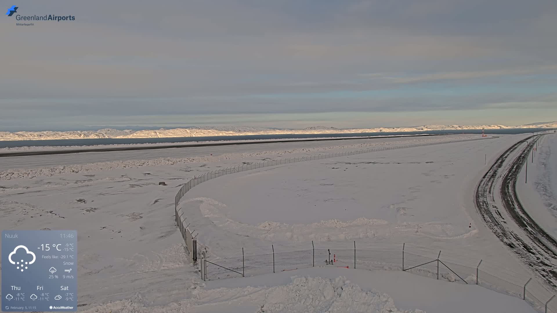 A snow-covered airfield and road stretch toward a distant, snow-capped mountain range under a partly cloudy sky.