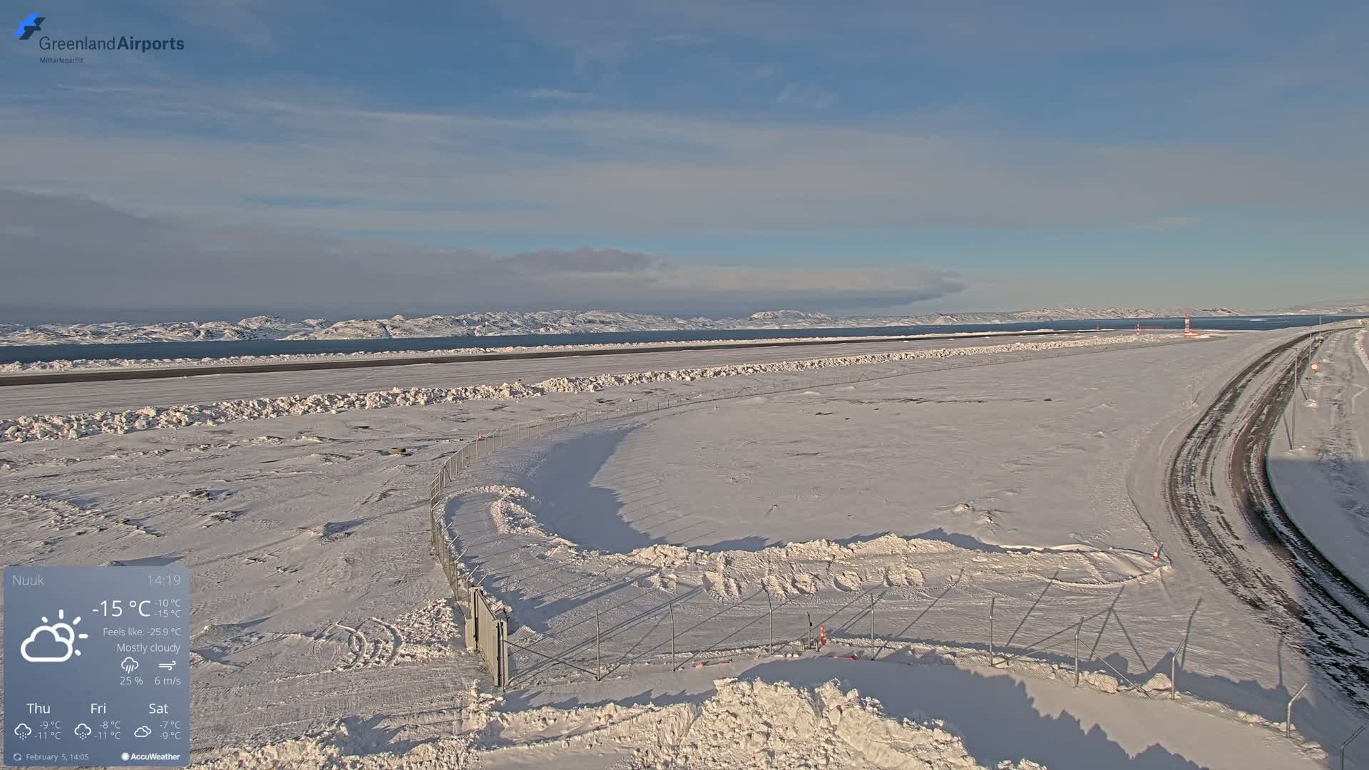 A snow-covered airport runway and surrounding area extend to a body of water with distant mountains under mostly cloudy skies.