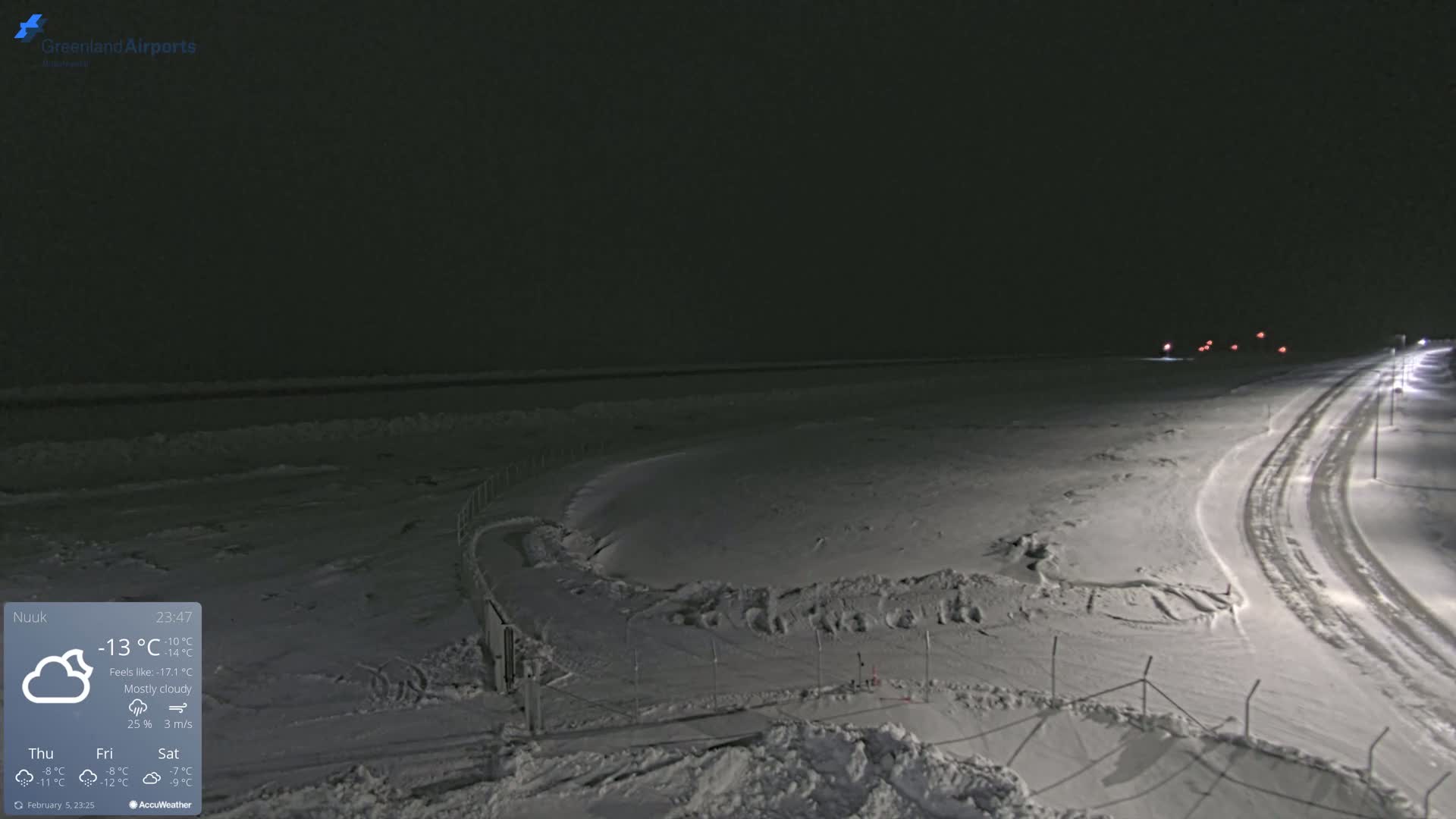 A snow-covered, nighttime landscape shows a road curving through a fenced-in area, with mostly cloudy skies and a temperature of -13°C.
