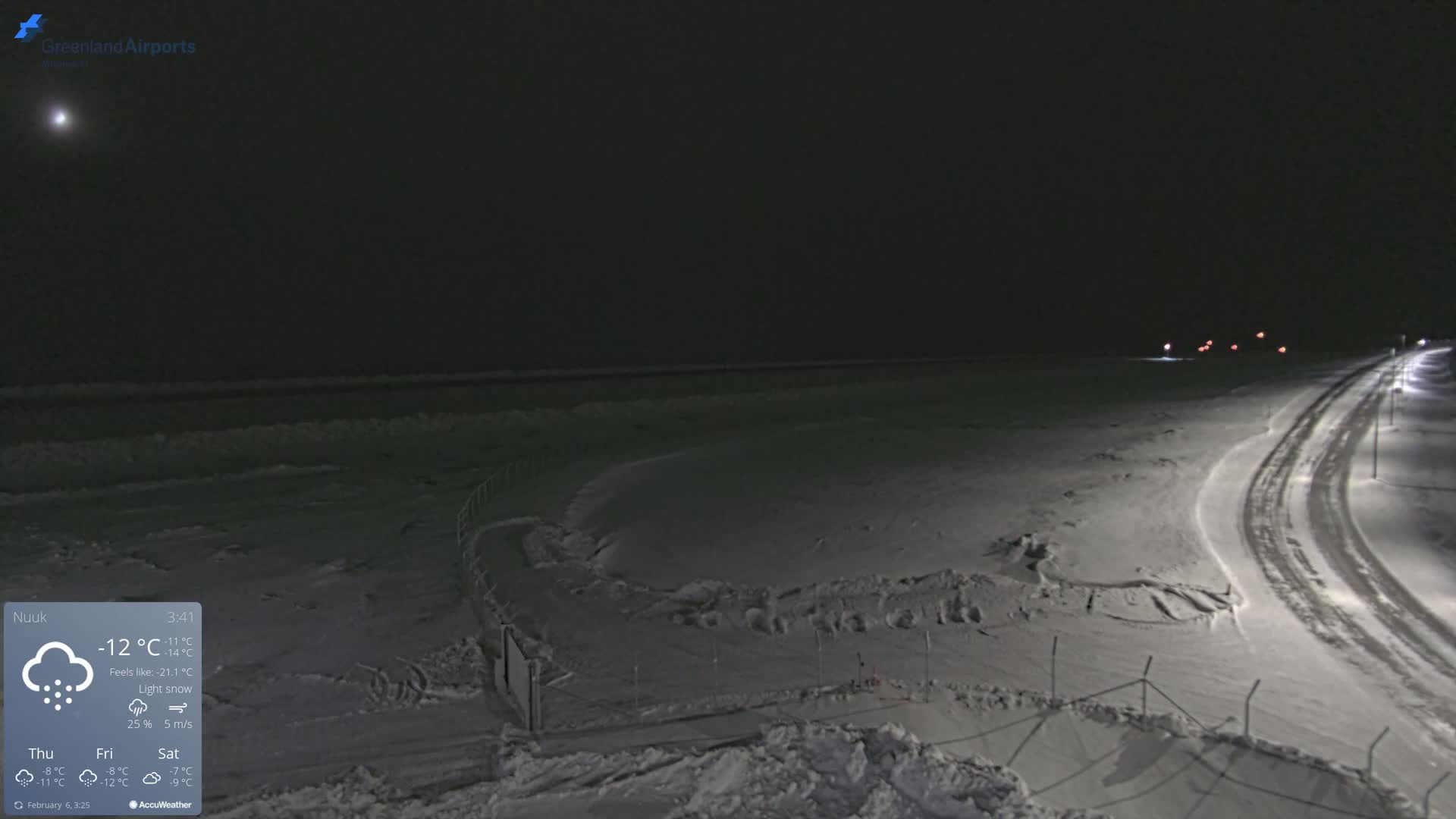 A snow-covered landscape at night, under a lightly snowing sky, shows a road with tire tracks curving towards a dimly lit area beyond a fence.