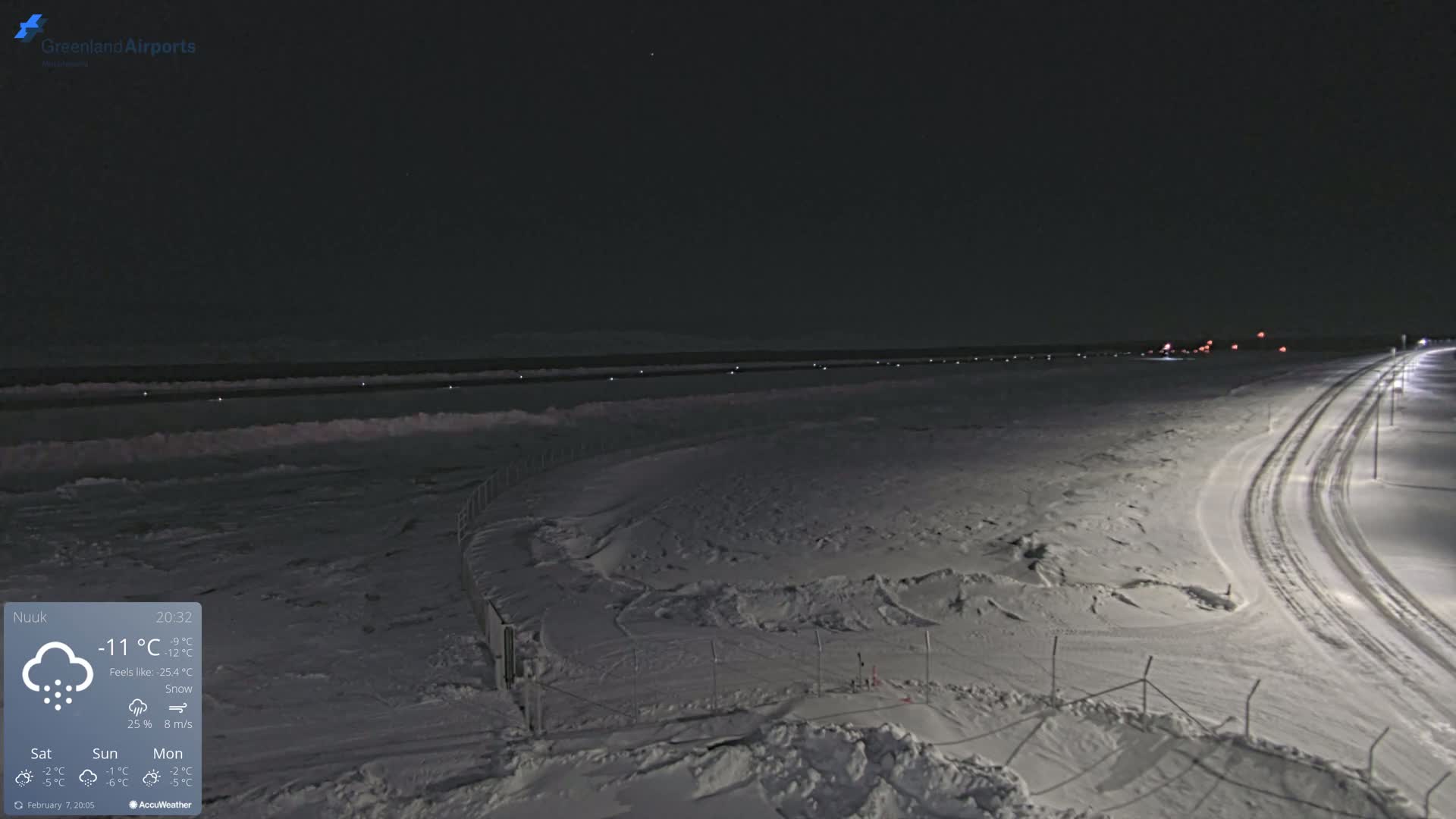 A snow-covered, nighttime landscape shows a roadway curving toward a body of water under a dark sky with light snow falling.