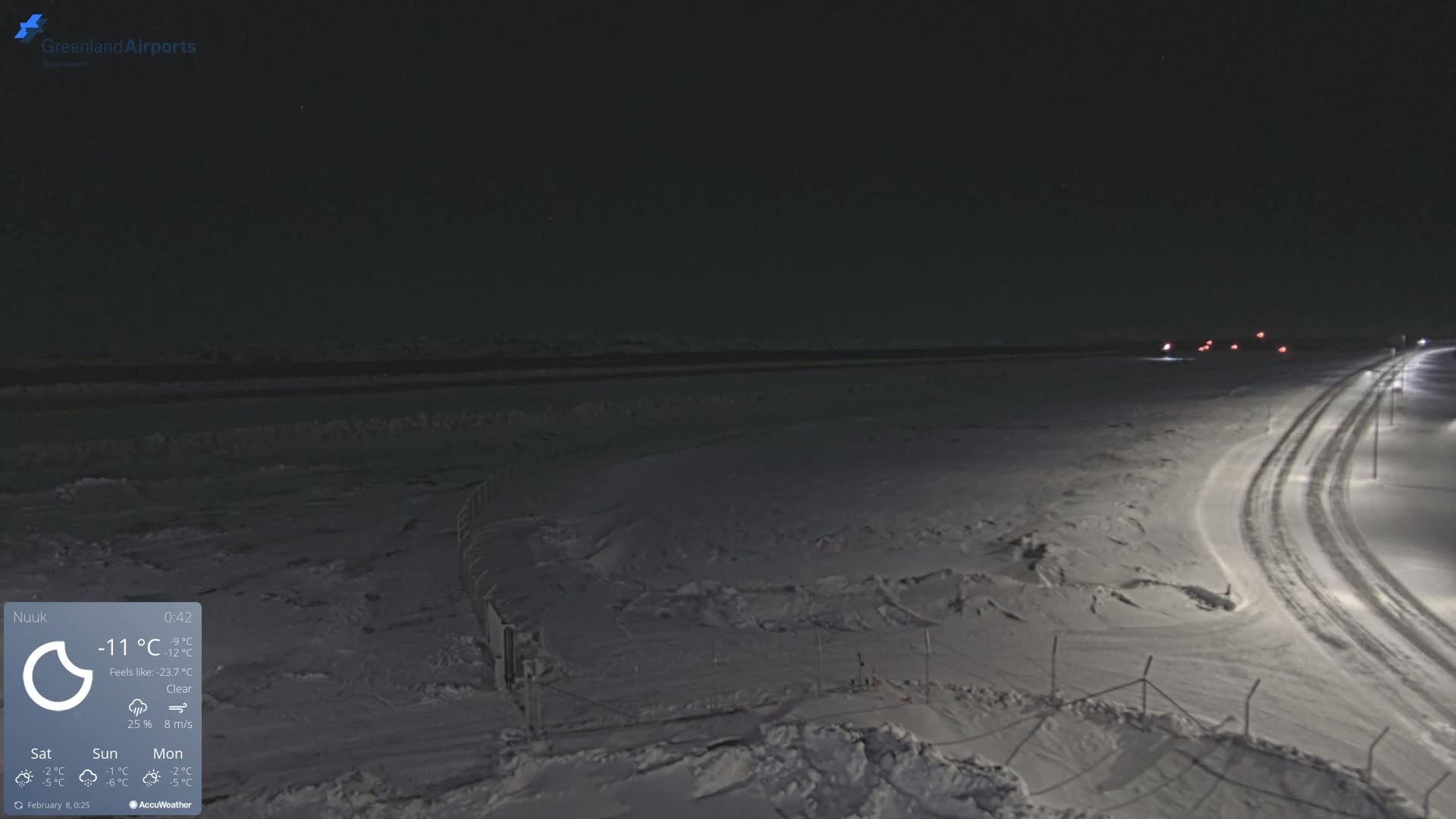 A snow-covered landscape at night under a clear sky, showing a road with tire tracks and distant lights.