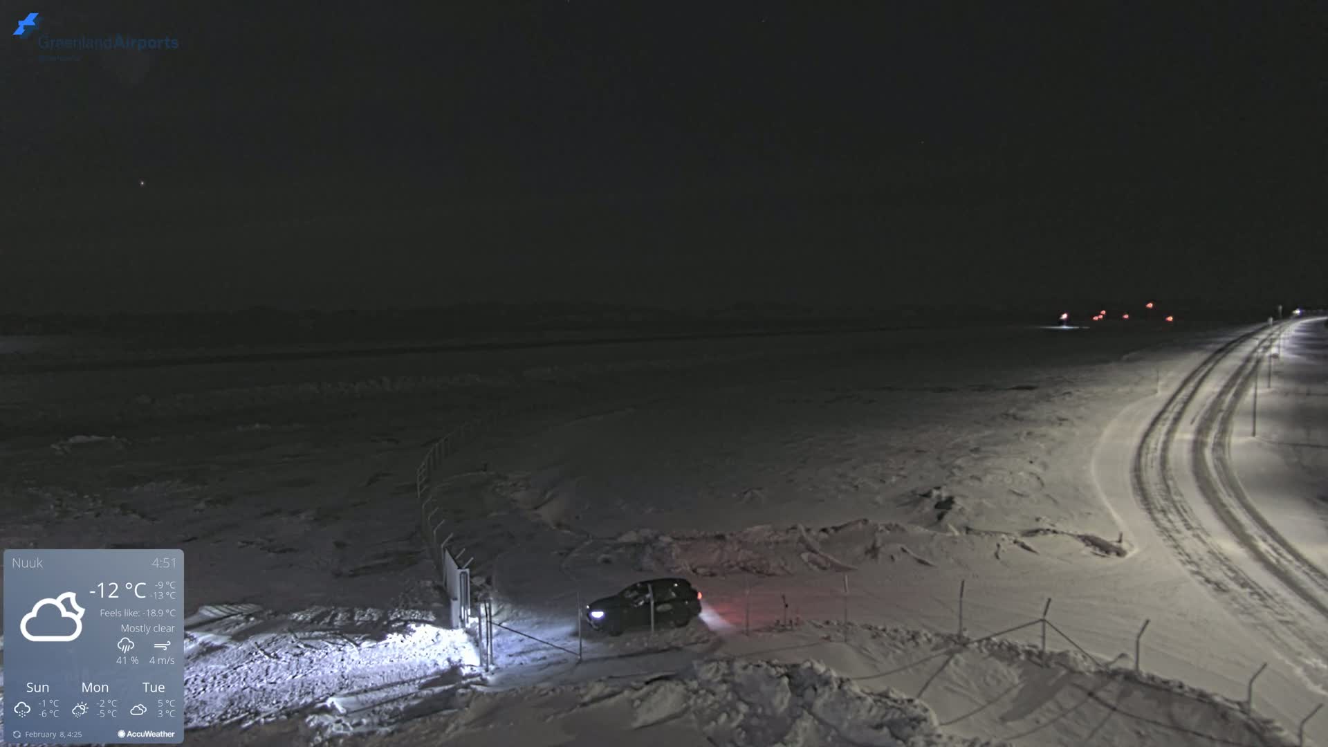 A dark, snow-covered landscape at night, under mostly clear skies, shows a single car near a fence and a road with faint vehicle lights in the distance.