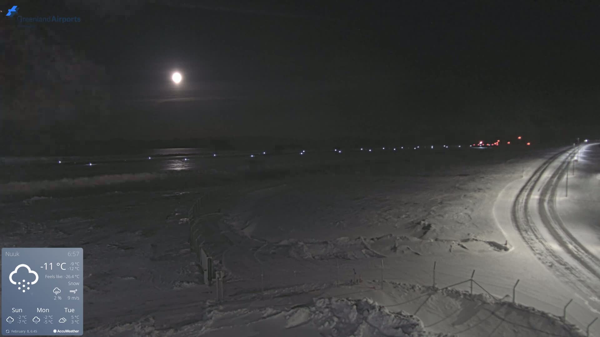 A bright moon illuminates a snow-covered landscape at night, with a road and a line of lights visible in the distance under snowy conditions.