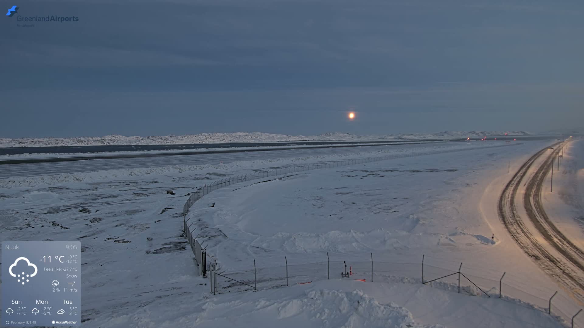 A snow-covered airport runway and road are visible under a twilight sky with a bright moon, and light snow is falling.