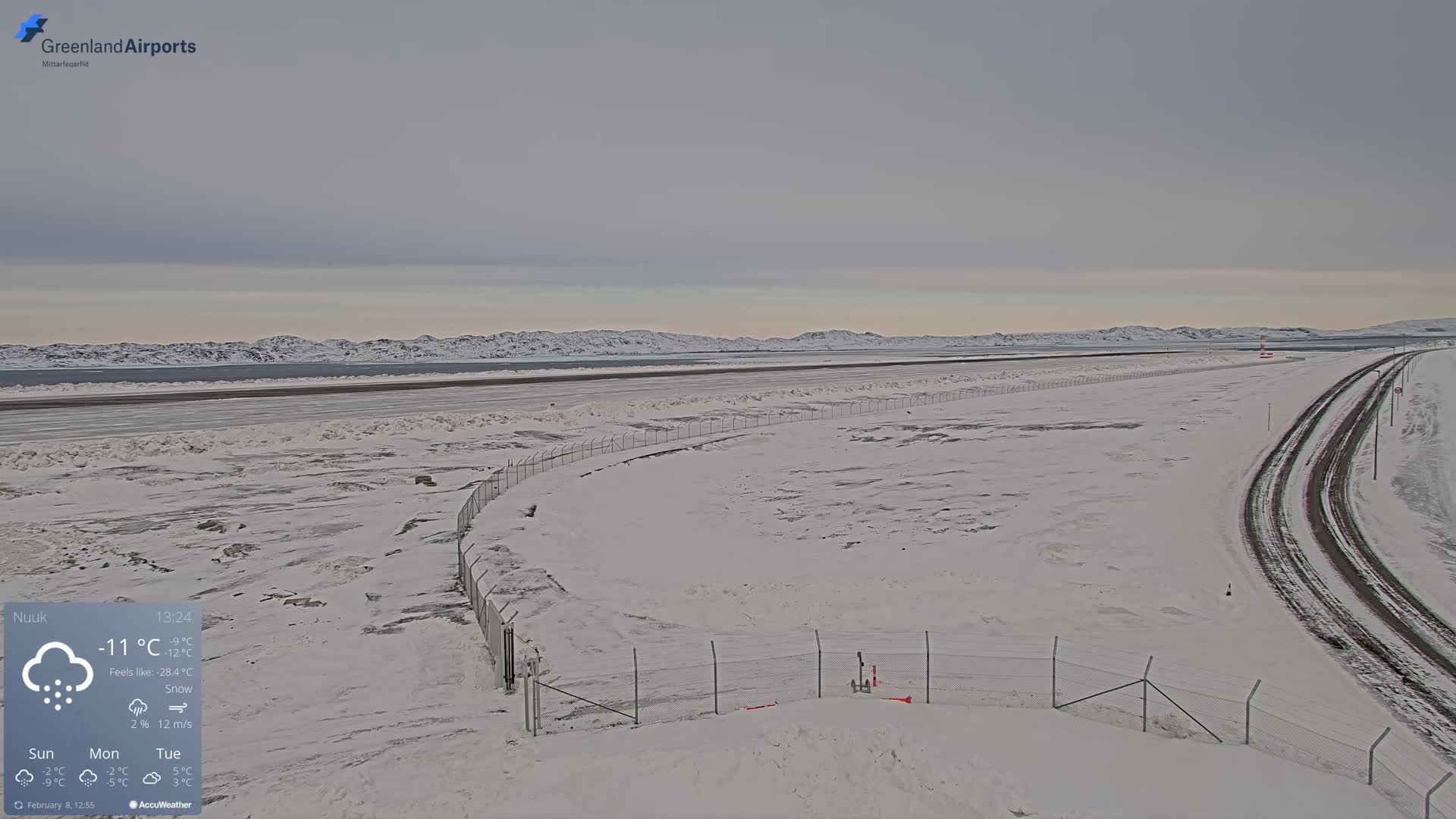 A snow-covered airport runway and surrounding area extend to a distant, snow-capped mountain range under a cloudy sky.