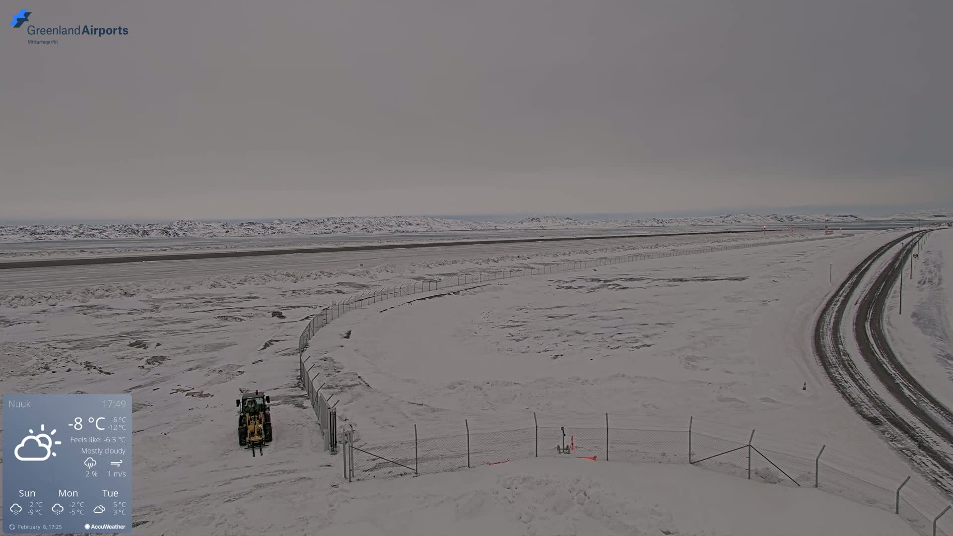 A mostly cloudy, snowy landscape features a snow-covered airfield, a road, and a small tractor near a chain-link fence.