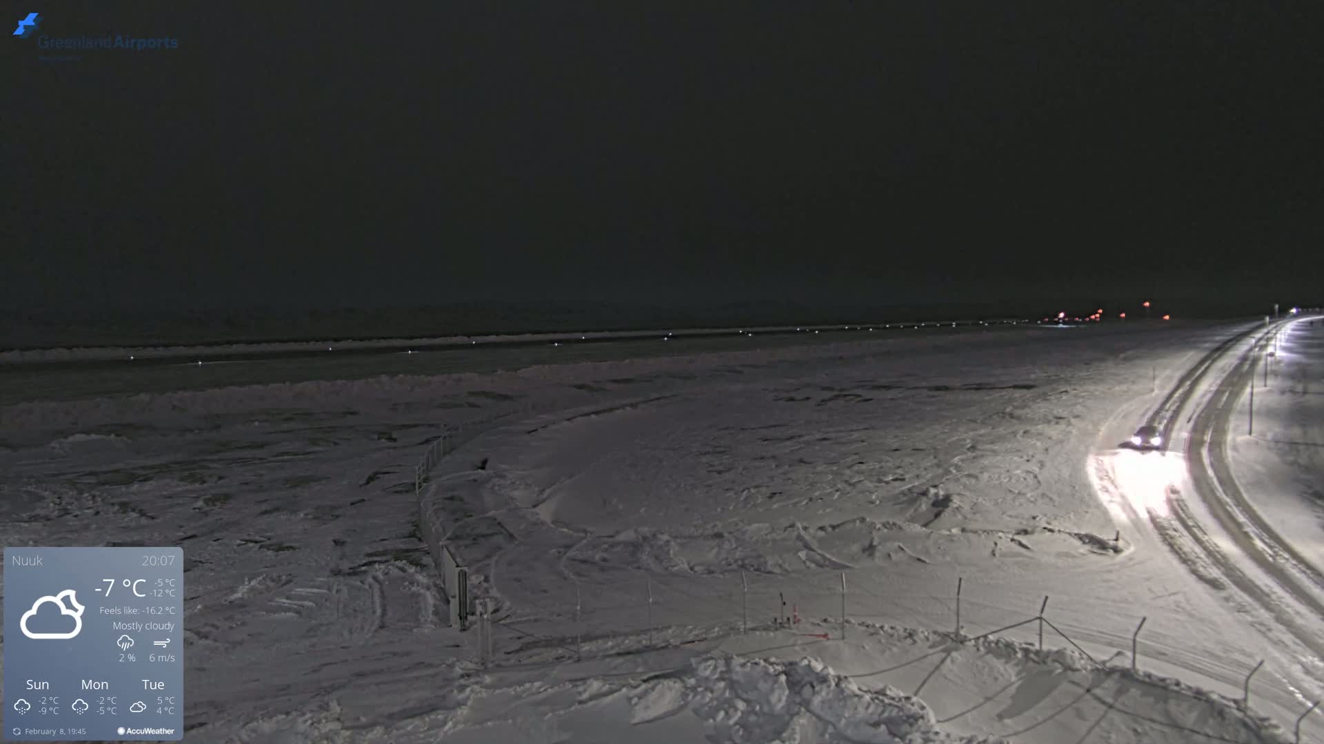 A mostly cloudy night scene shows a snow-covered airfield with a vehicle driving on a nearby road.