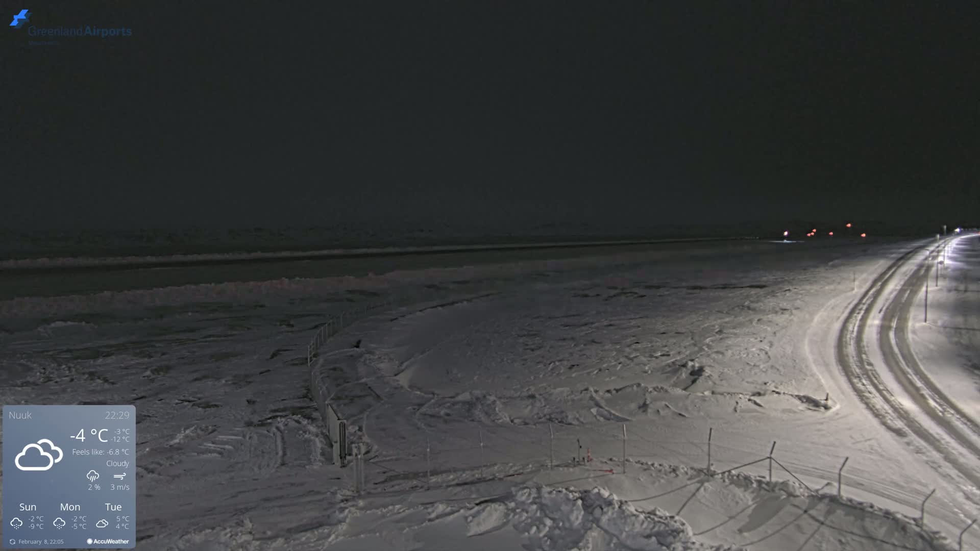A snow-covered landscape at night, illuminated by lights along a road, with a body of water visible in the distance under a cloudy sky.