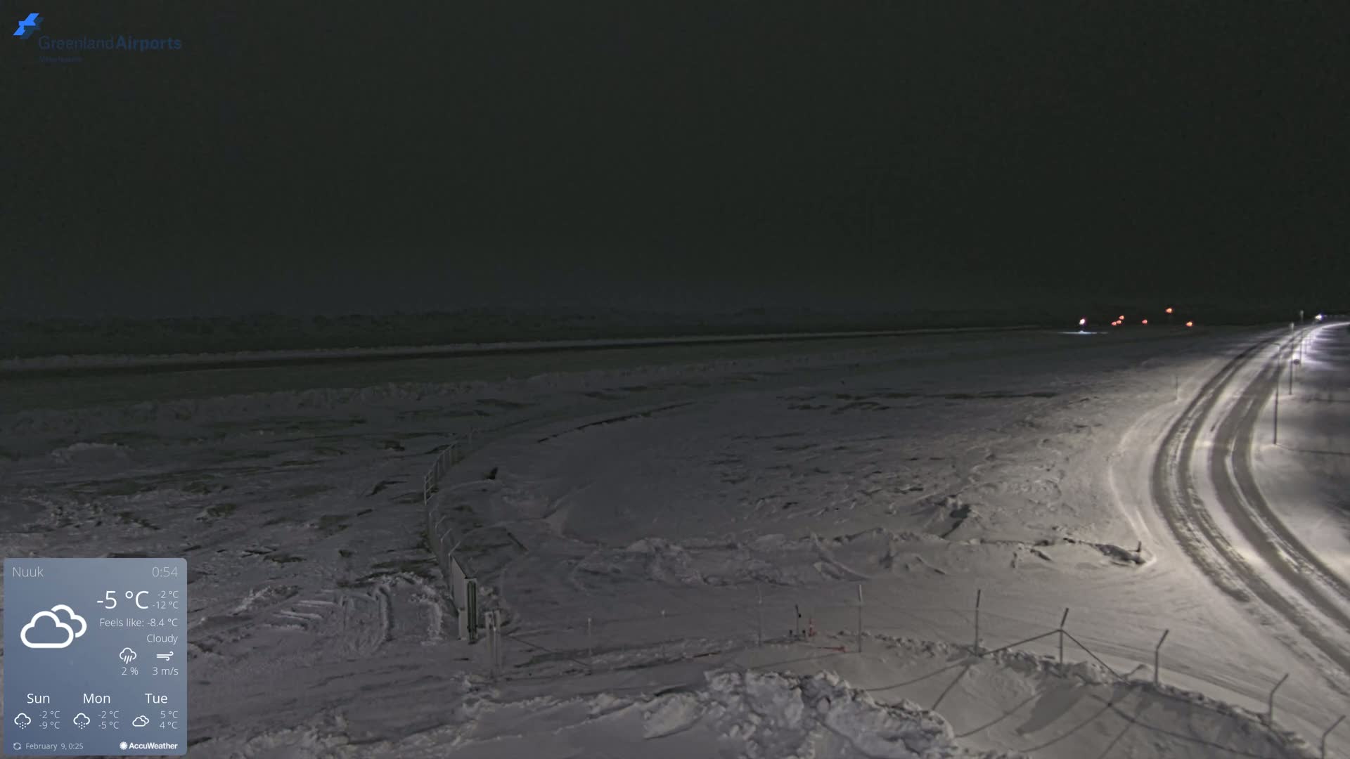 A snow-covered landscape at night is illuminated by streetlights along a road, with a cloudy sky.