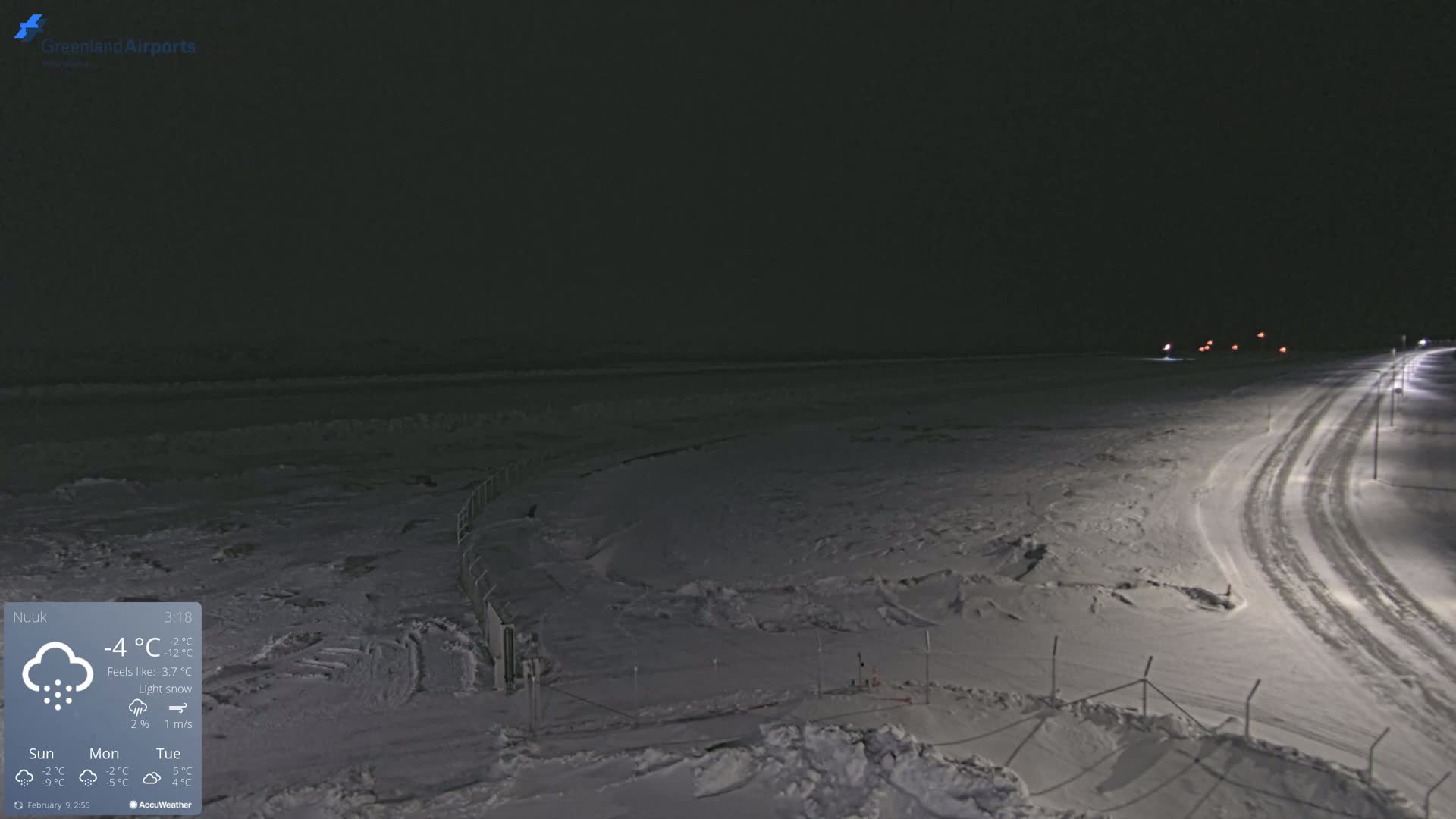 A snow-covered landscape at night under light snowfall, showing a road with tire tracks and faint lights in the distance.
