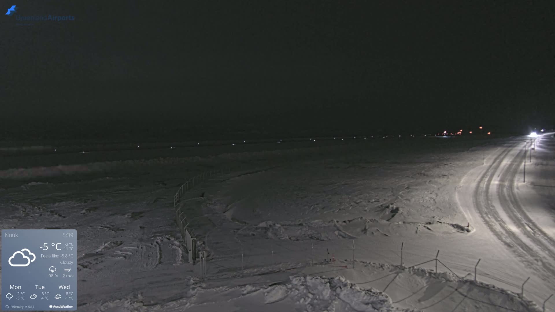 A snow-covered landscape at night, with a road and a body of water in the distance under a cloudy sky.